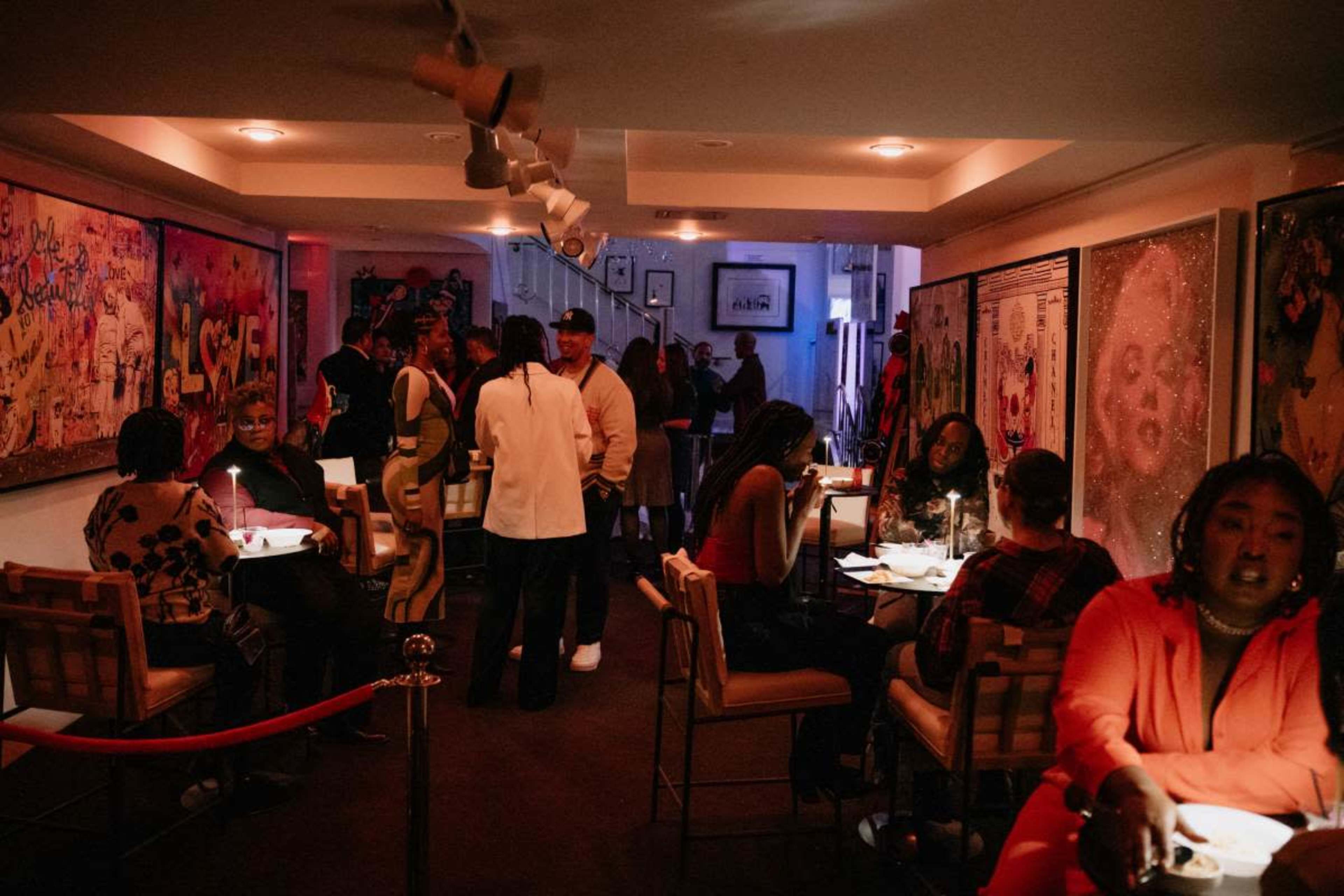 A lively indoor space with people socializing at tables and admiring art on the walls, illuminated by soft lighting.