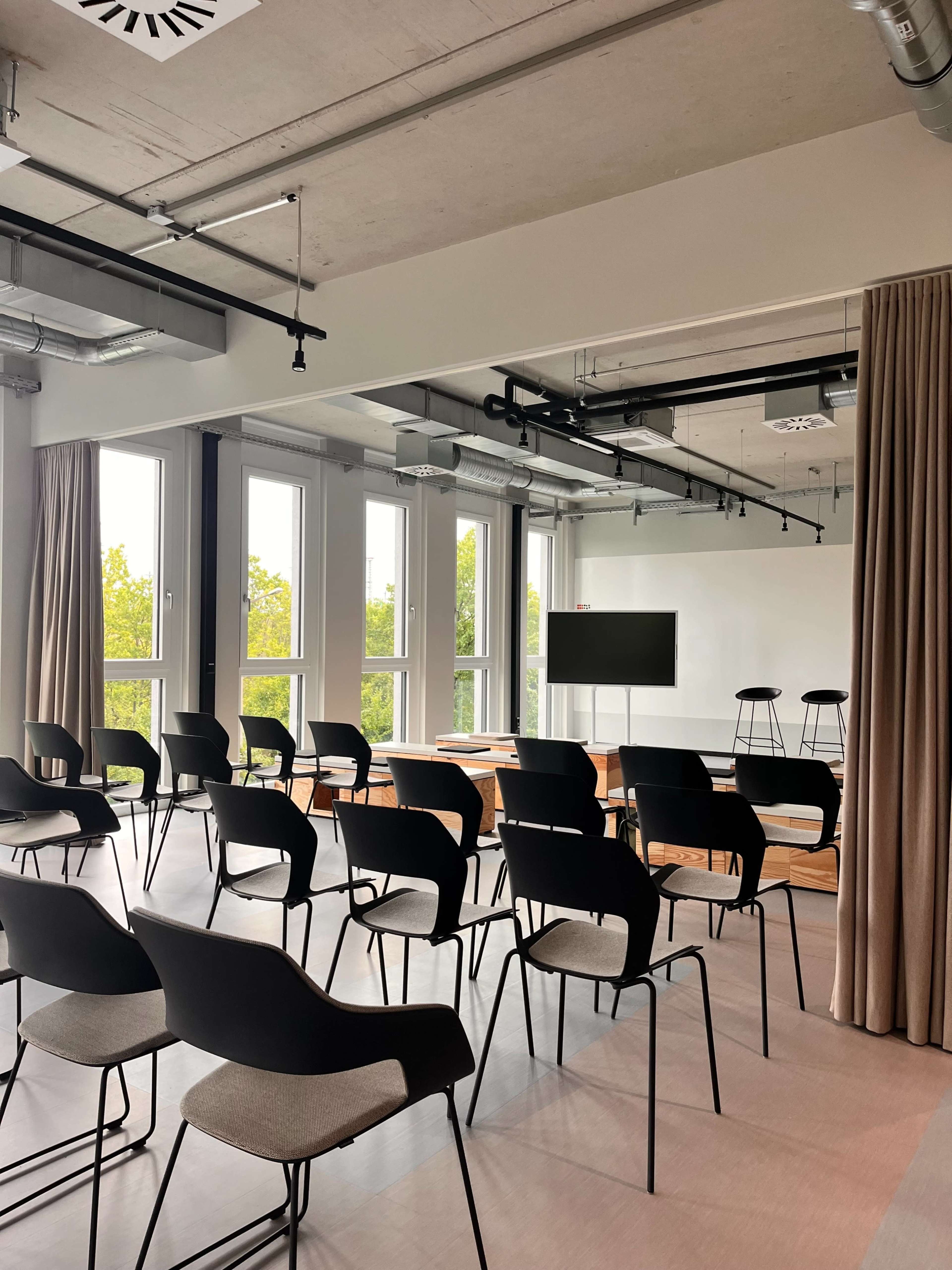 The image shows a modern conference room set up with rows of black chairs facing a presentation screen, alongside tables and large windows allowing natural light.