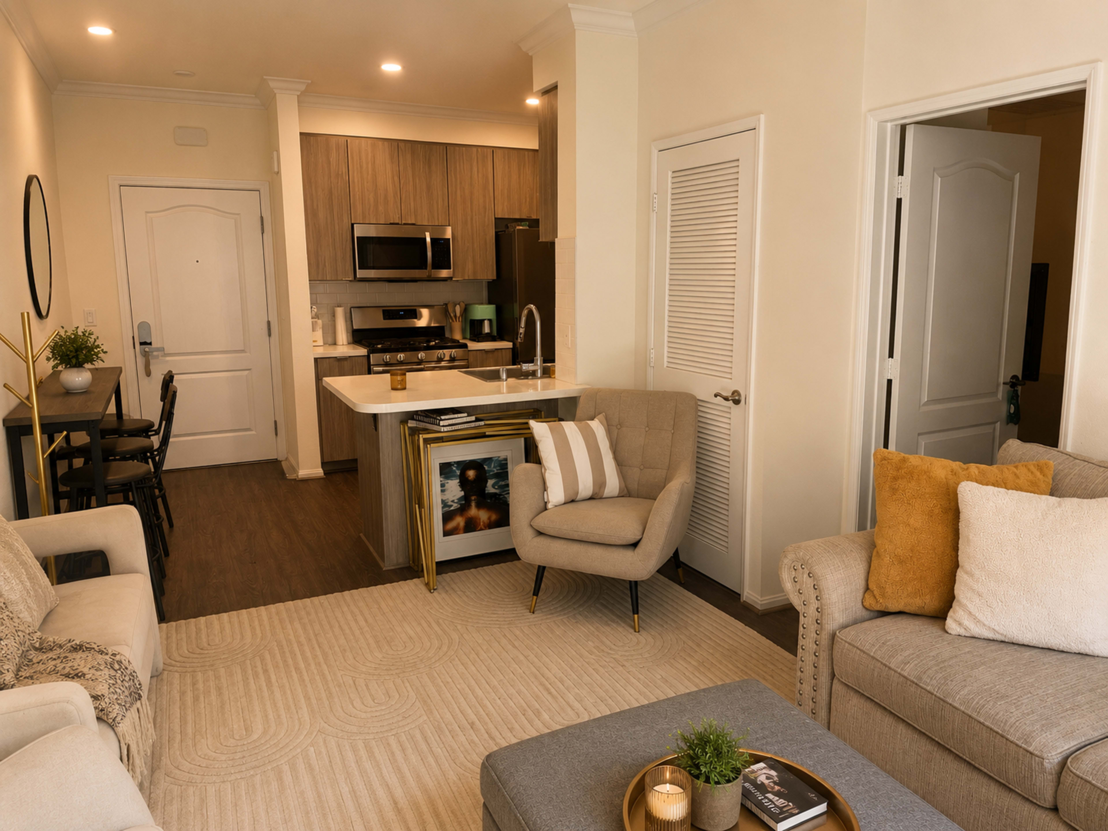 The image shows a modern living room and kitchen area in an apartment, featuring a sofa, armchair, and dining table with a kitchenette in the background.
