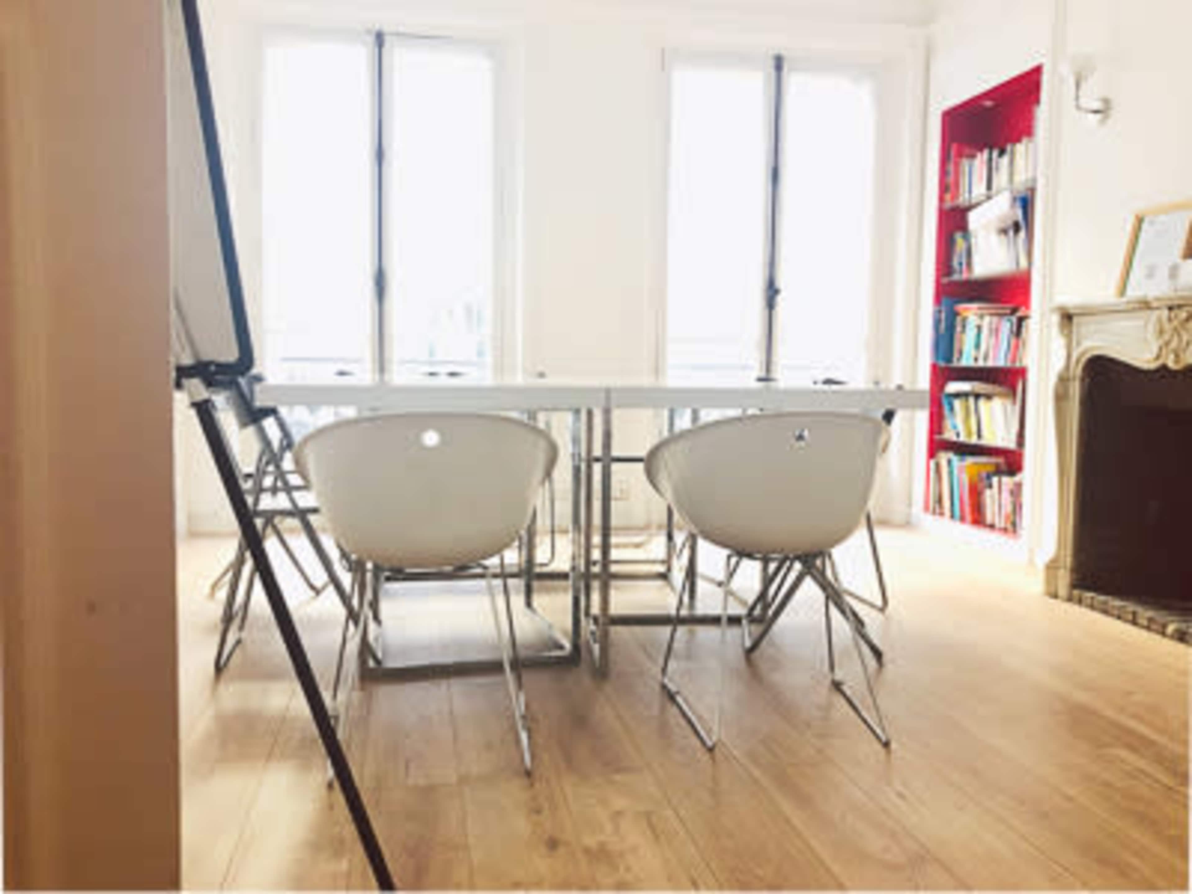 A bright meeting room features a rectangular table surrounded by four white chairs, with a bookshelf filled with colored books in the background.
