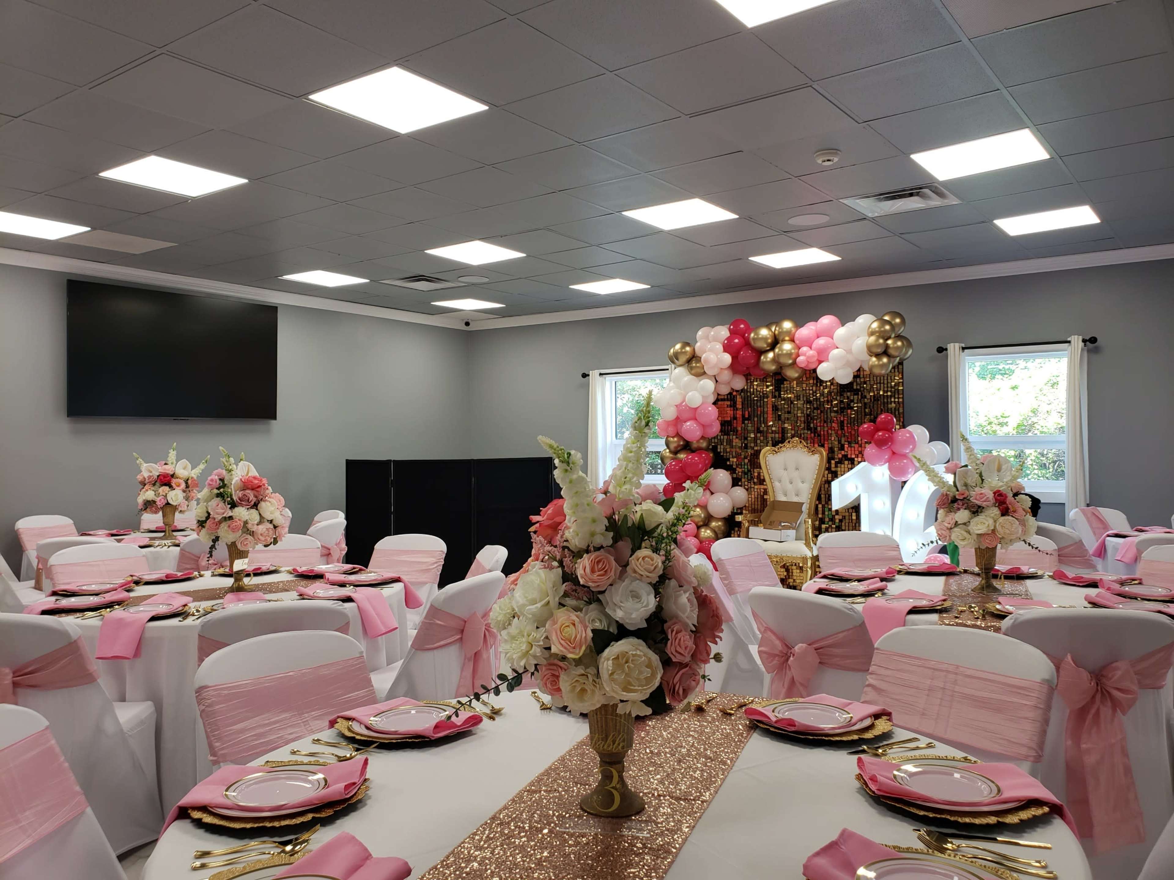 The image shows a elegantly decorated banquet hall with tables set with white tablecloths and pink accents, featuring floral centerpieces and a backdrop of balloons.