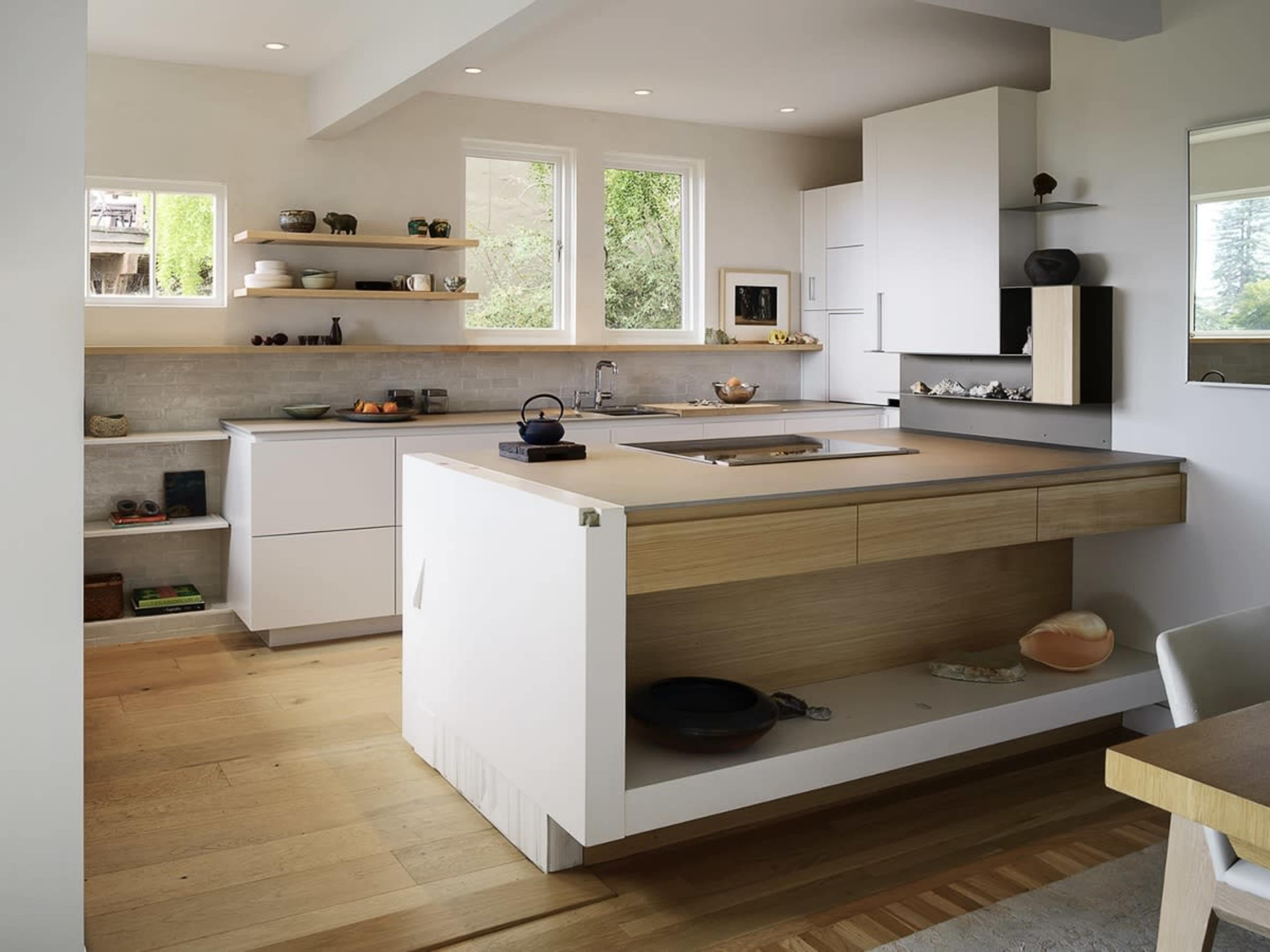 The image shows a modern kitchen with sleek white cabinetry, a central island featuring a wooden countertop, and natural light streaming through large windows.