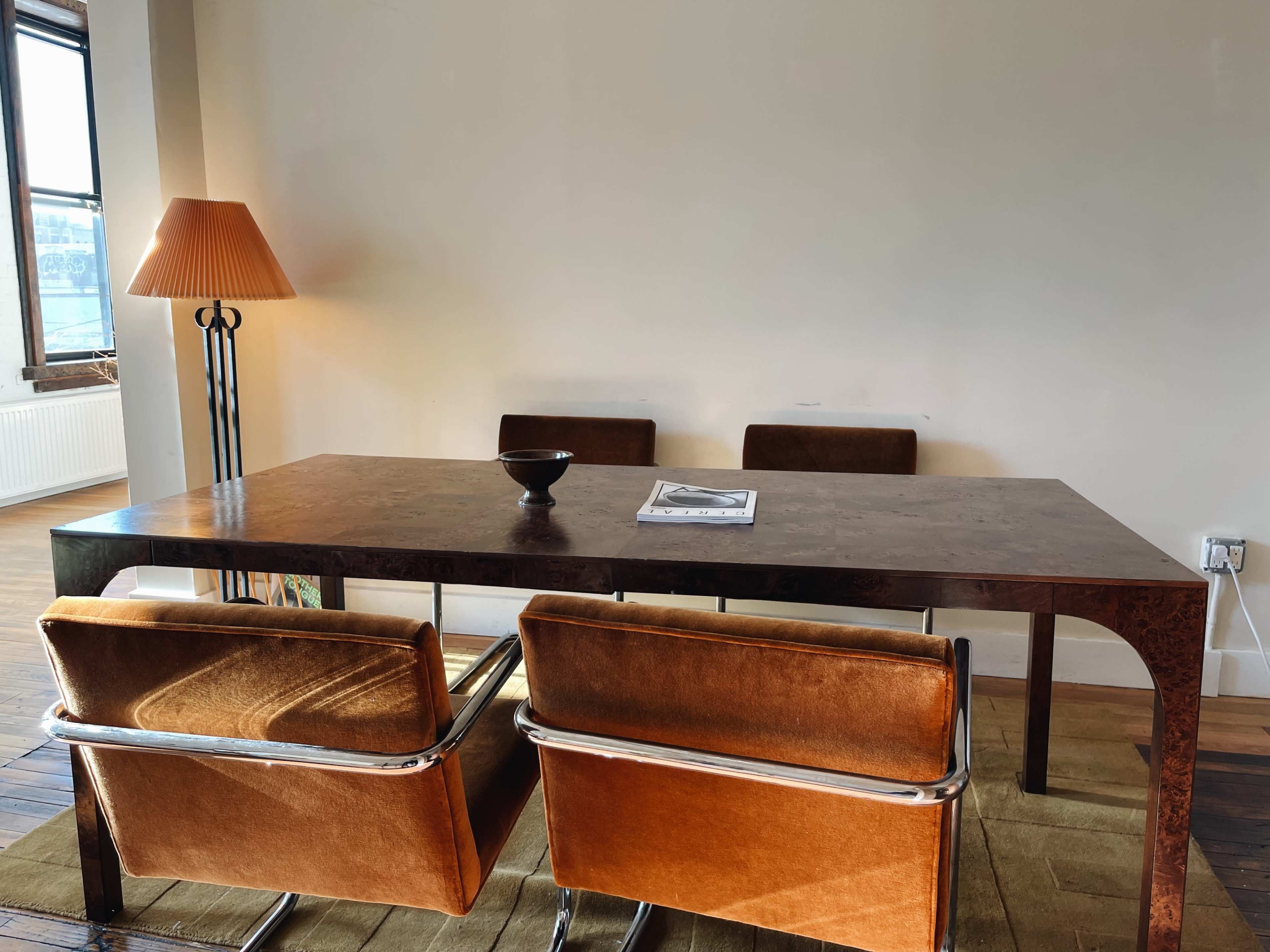 A rectangular wooden table is positioned in a sparsely furnished room, accompanied by four brown chairs and a standing lamp.