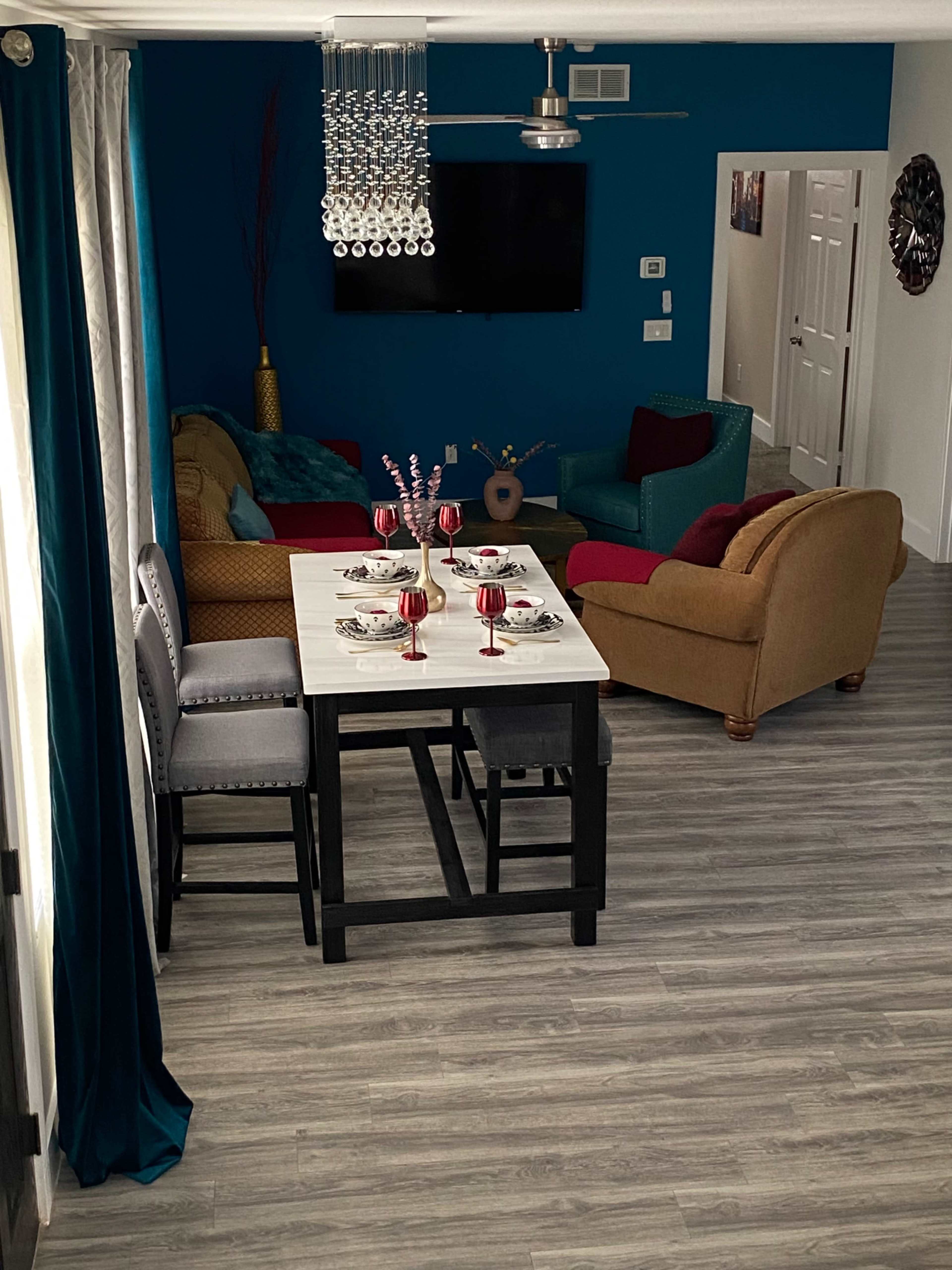The image shows a modern living and dining area featuring a table set for four, a television on the wall, and colorful furniture arrangements.