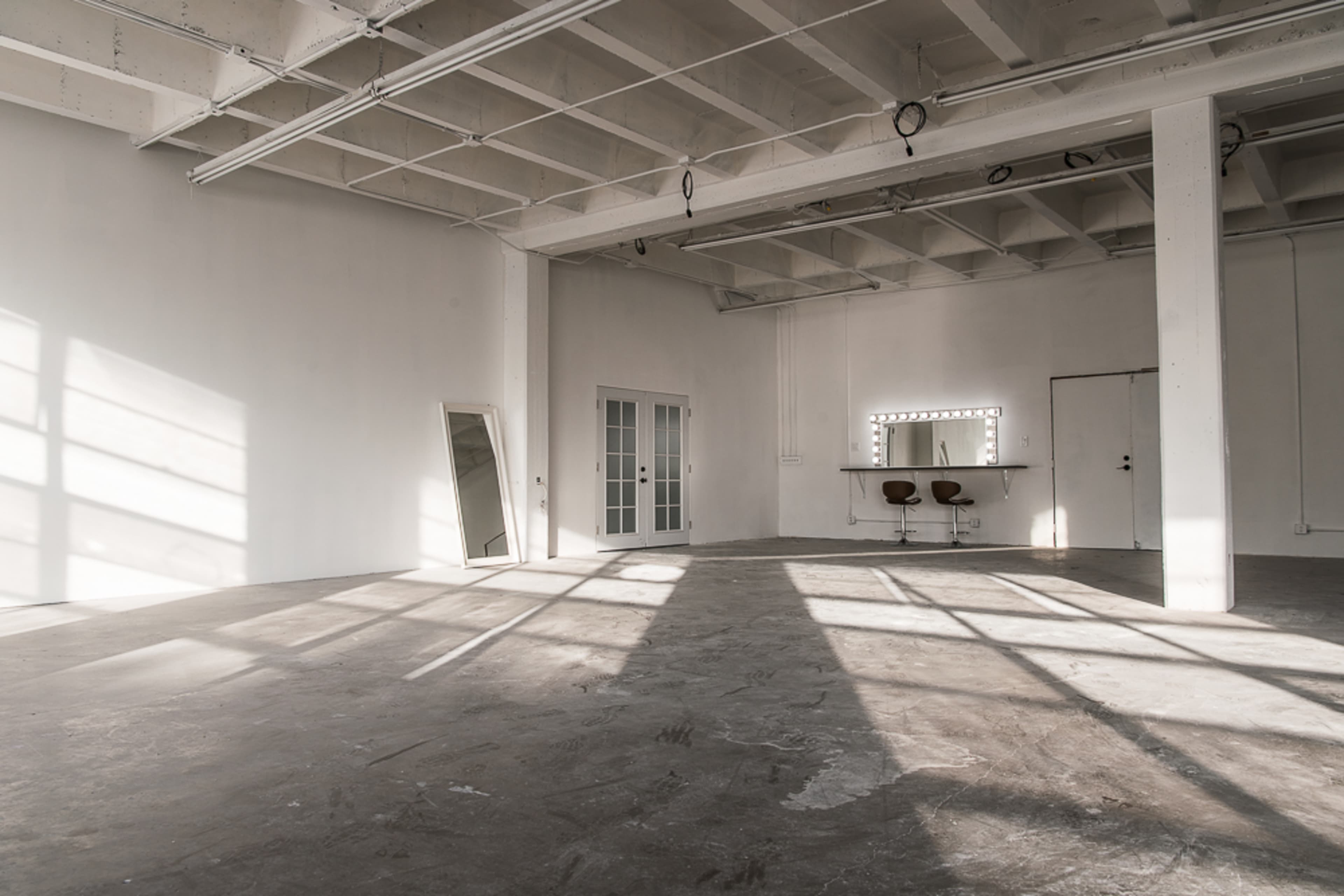 An empty room with a concrete floor, large windows casting shadows, a mirror leaning against the wall, and a vanity with two chairs.