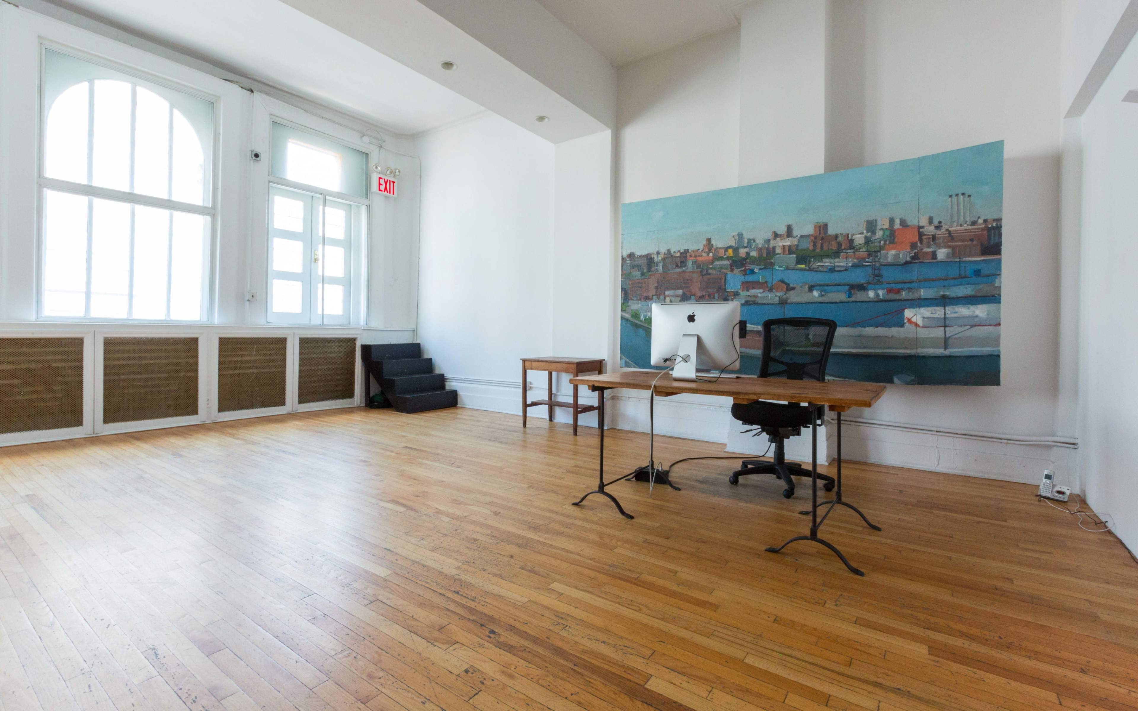 The image shows a minimalist office space with a wooden desk, an office chair, and a large painting on the wall, complemented by large windows letting in natural light.