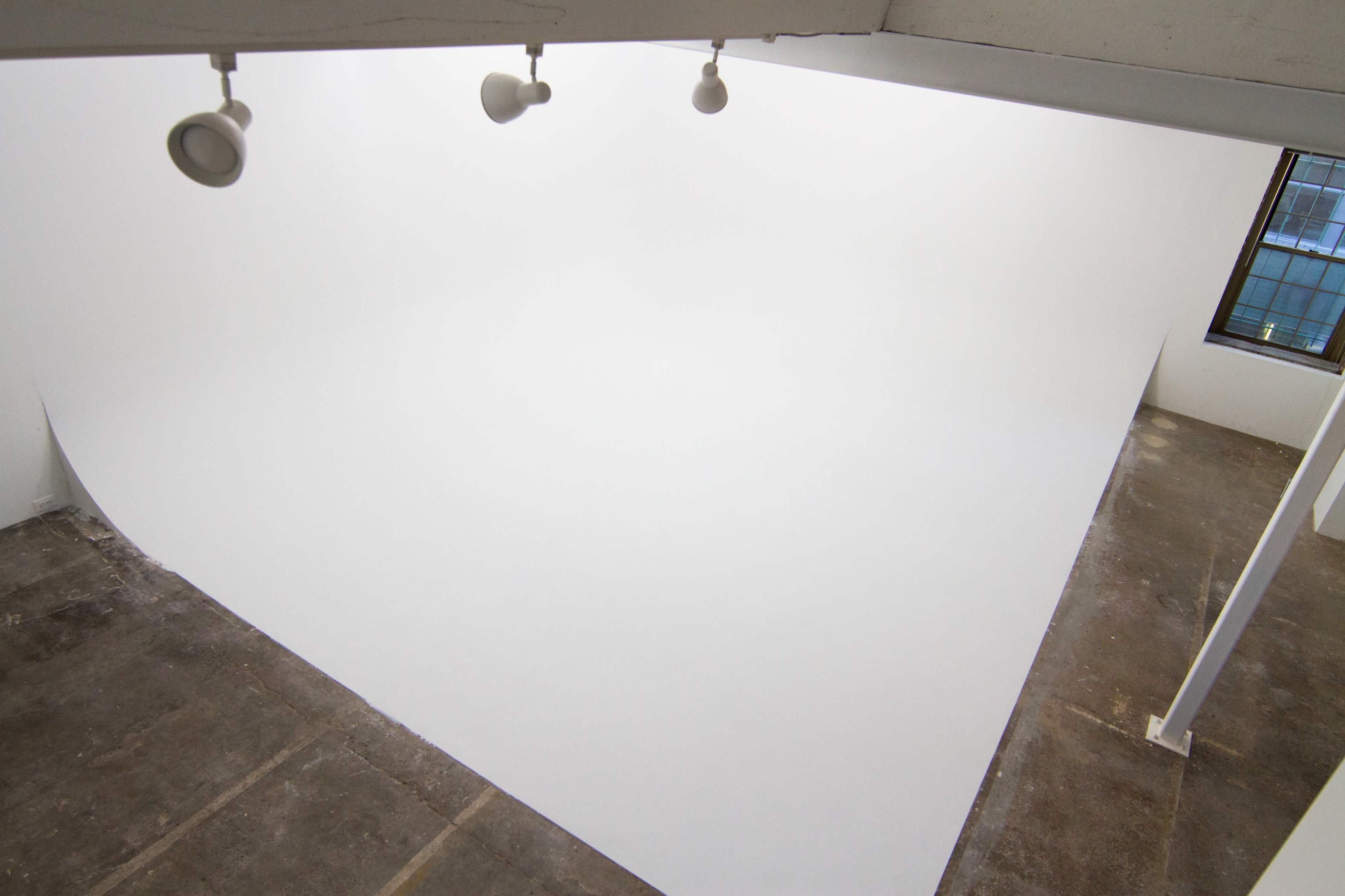 A spacious, empty room with a curved white wall and ceiling, illuminated by overhead spotlights.