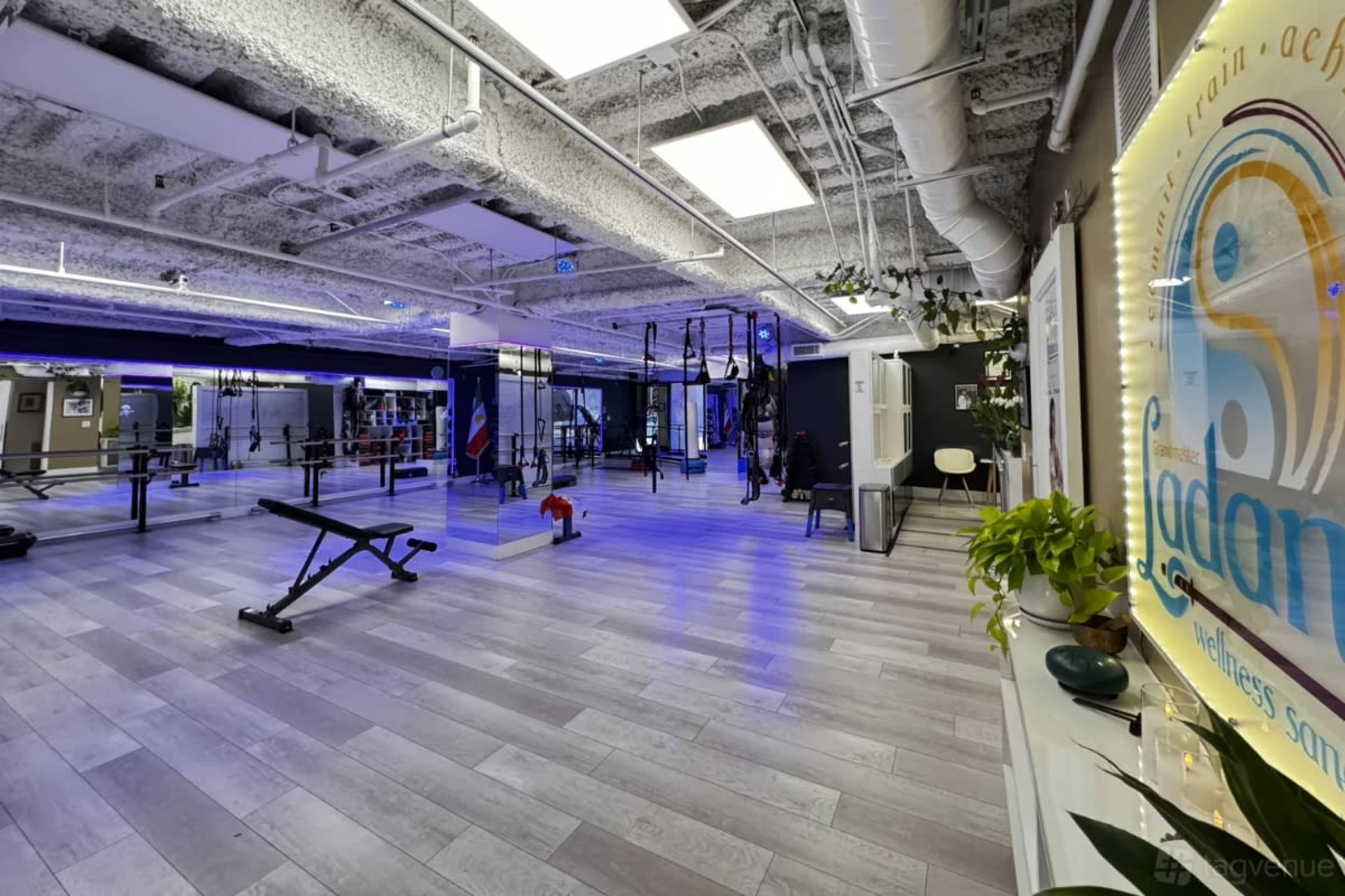 The image shows a modern fitness studio with various workout equipment, including weights and resistance bands, under bright lighting and a spacious layout.
