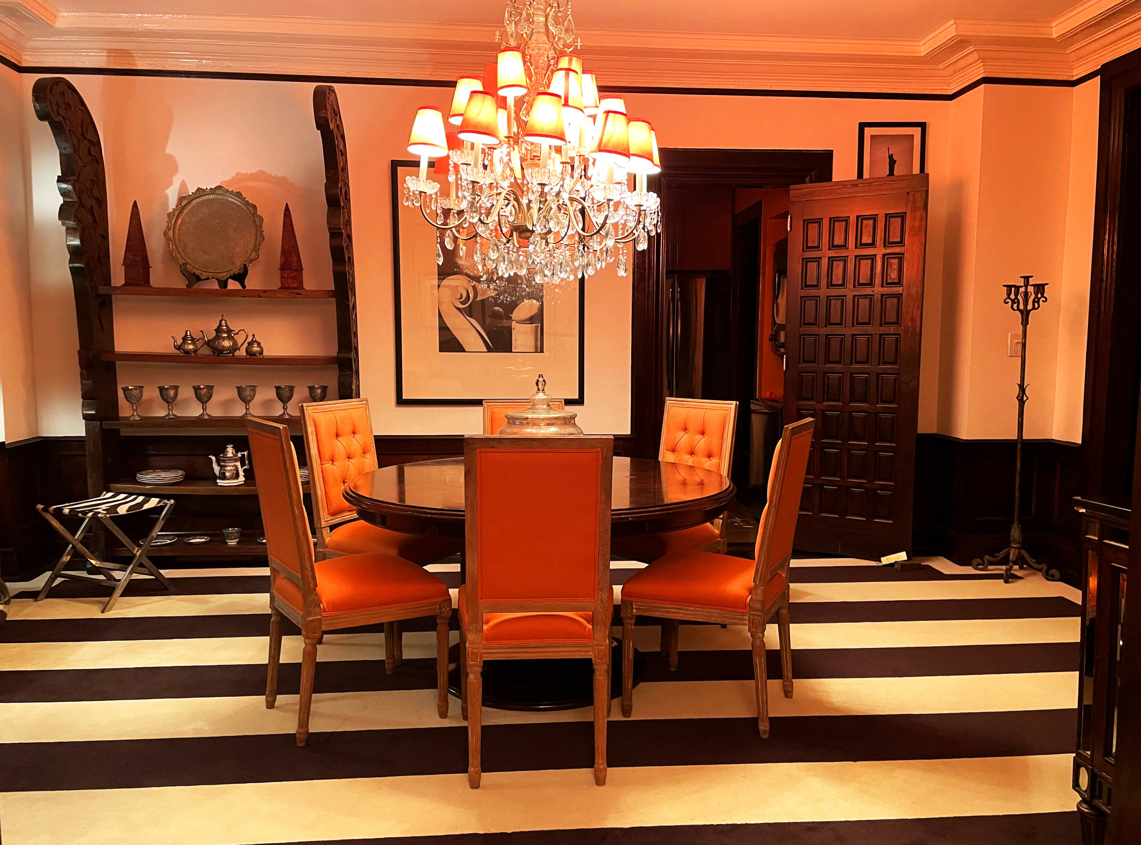 A circular wooden dining table surrounded by four orange upholstered chairs is illuminated by a chandelier in a room with striped flooring and dark wood accents.