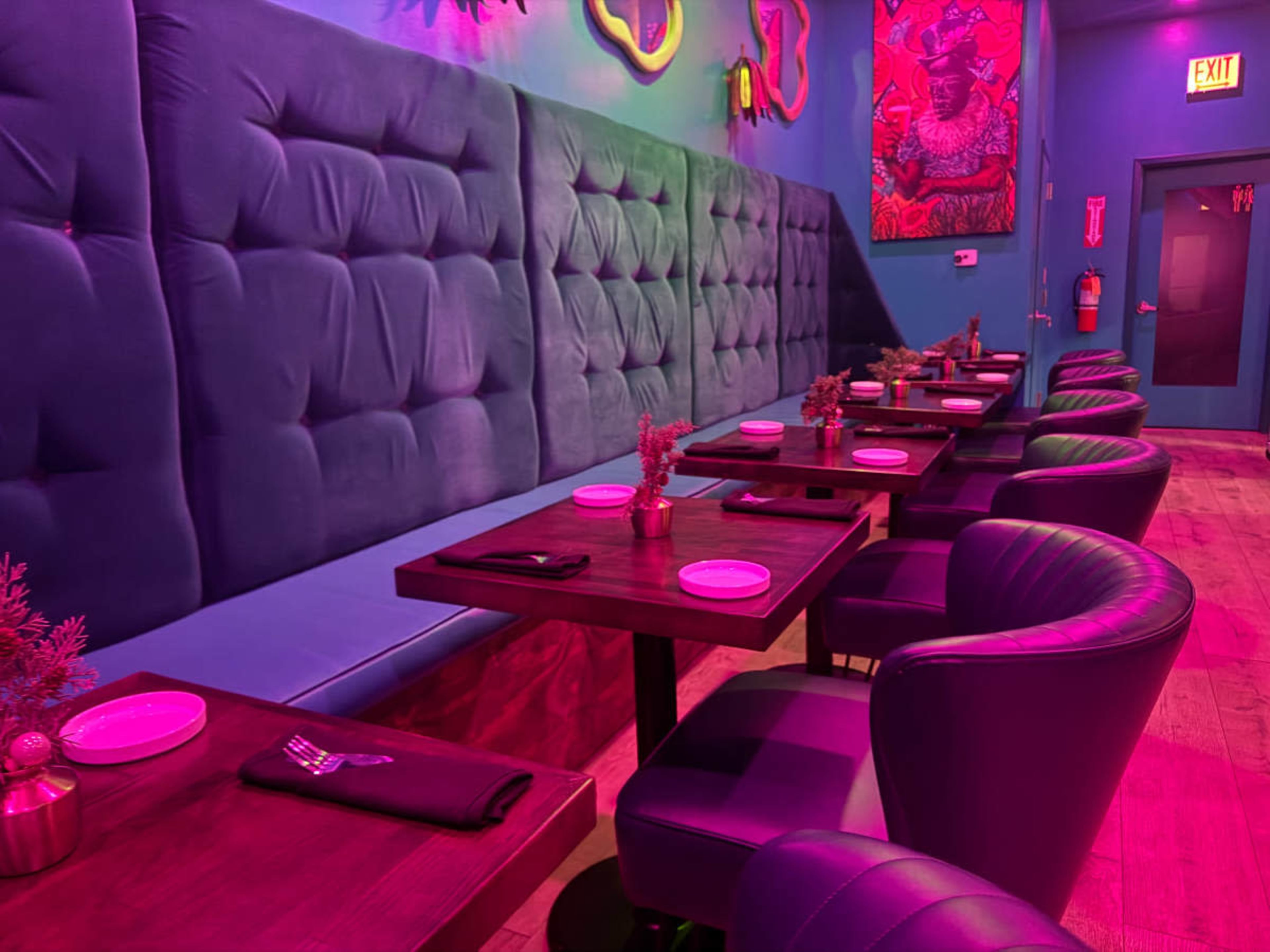 The image shows a restaurant interior with pink and purple lighting, featuring a row of tables and upholstered seating along a wall.