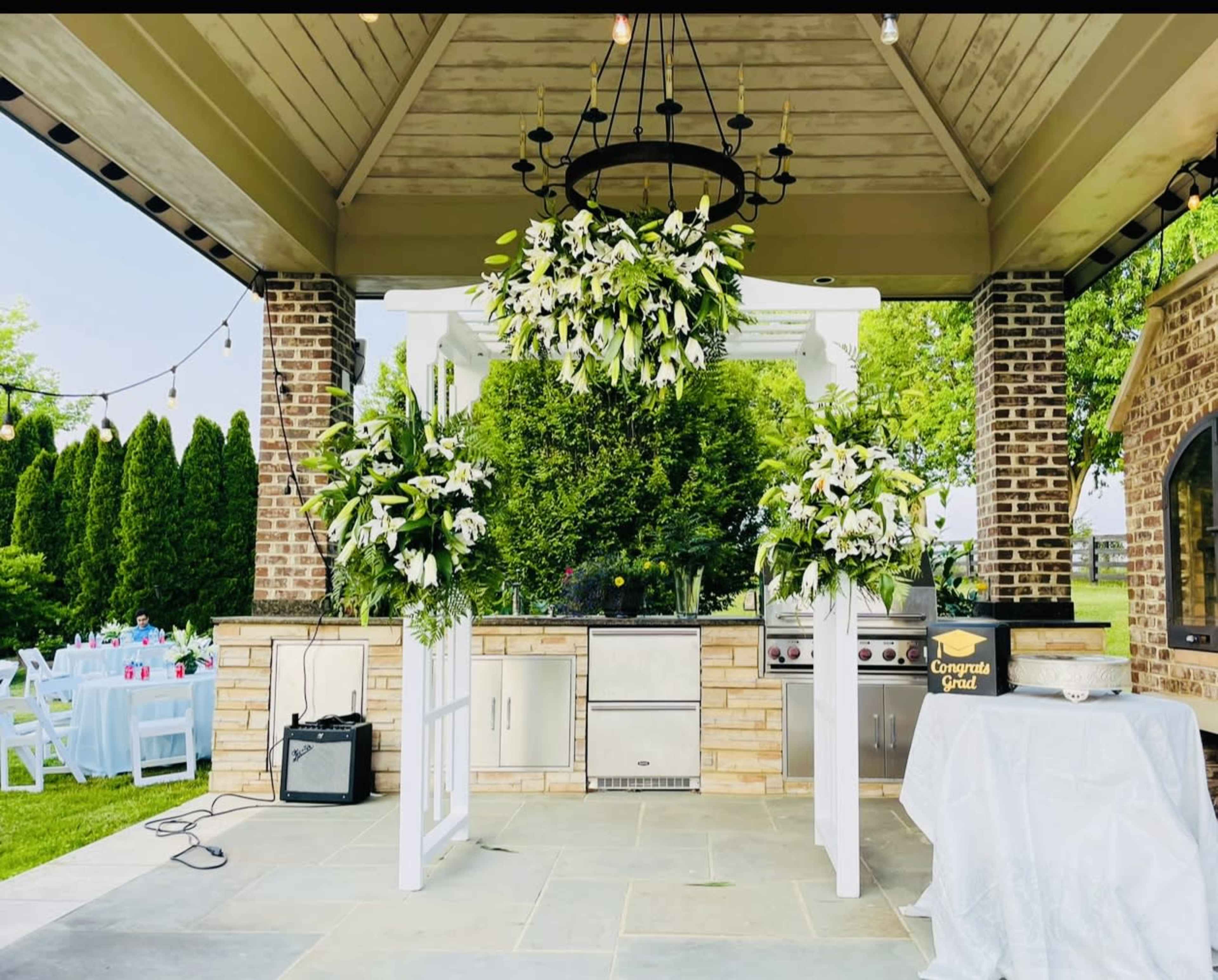The outdoor kitchen area features a stone grill, a large floral arrangement hanging from the ceiling, and two side arrangements on pedestals, all set under a covered structure.