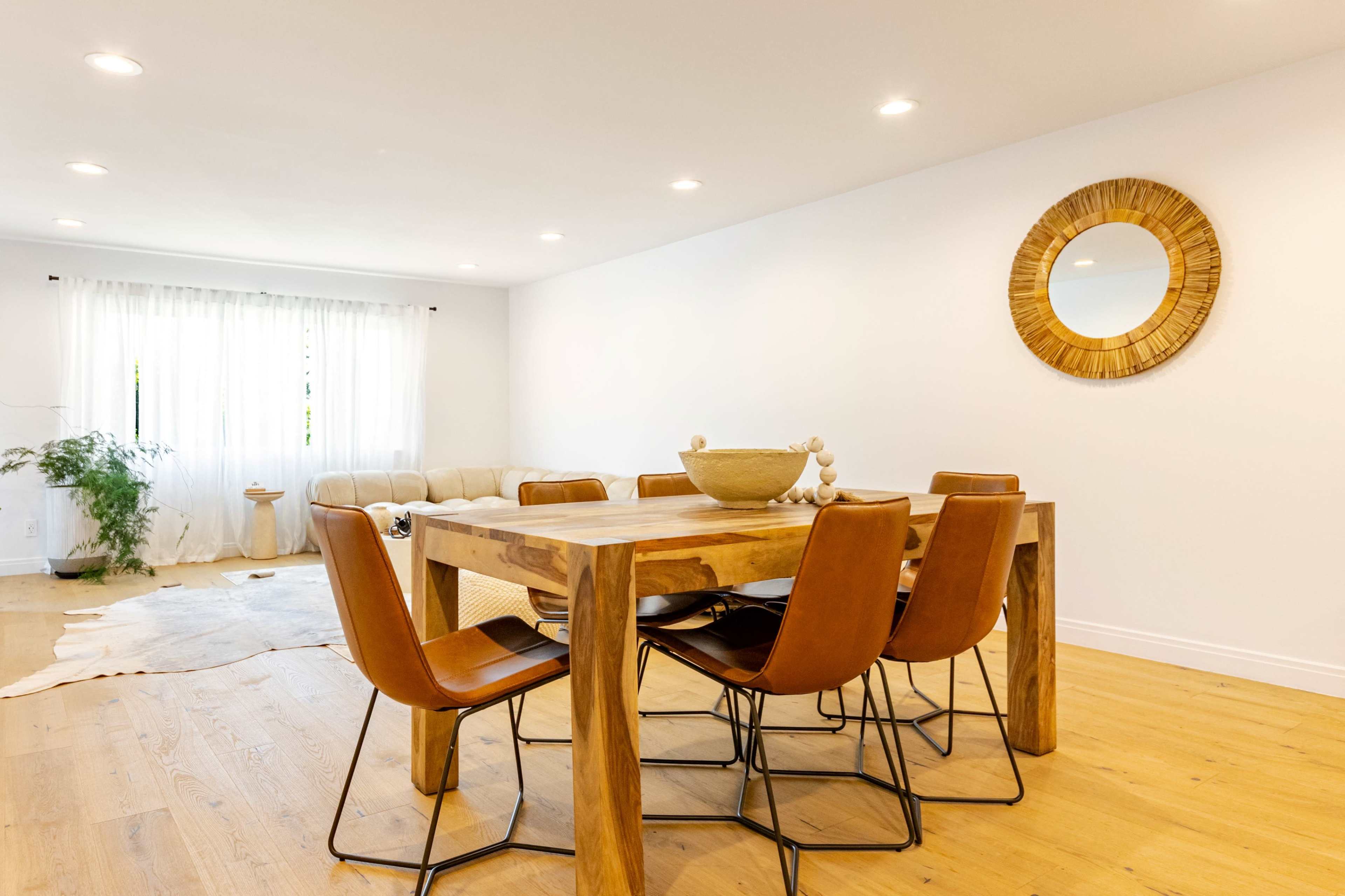 The image shows a modern dining room featuring a wooden table surrounded by brown chairs, with a light-colored rug and a mirror on the wall.