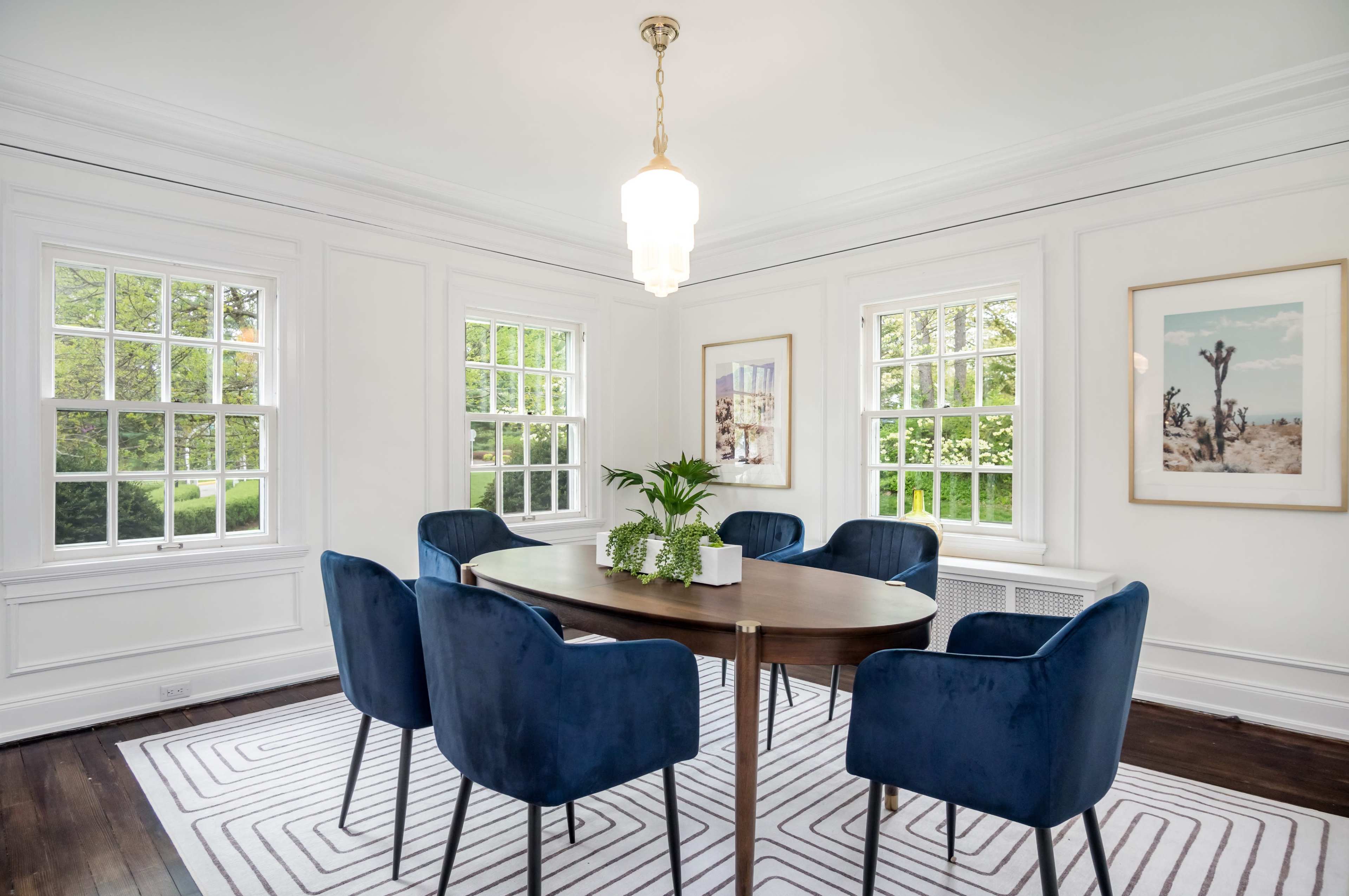 A dining room features a round wooden table surrounded by six blue upholstered chairs, with large windows providing natural light and plants as decor.