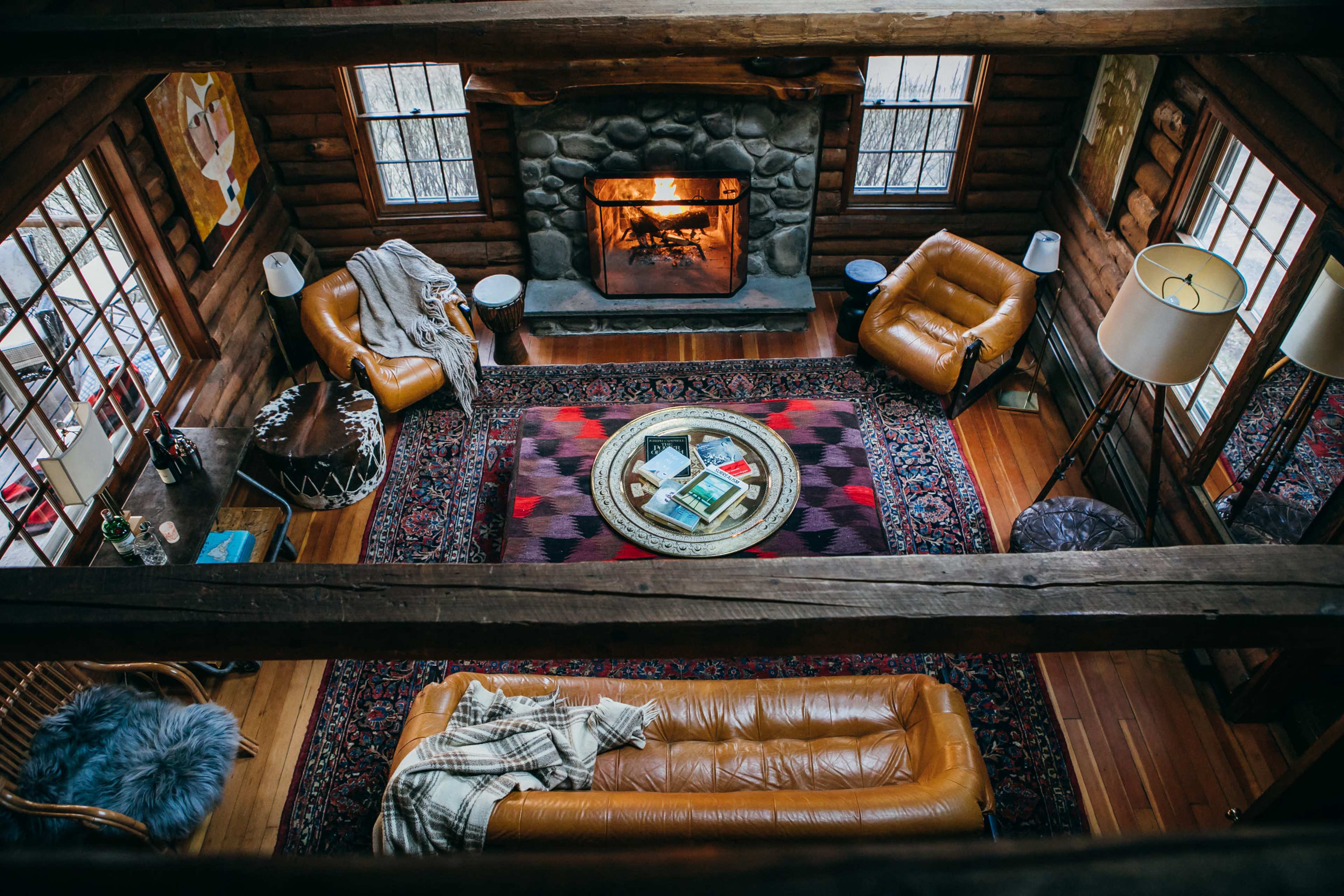 A cozy cabin living room features a stone fireplace, brown leather furniture, and a round coffee table surrounded by decorative rugs.
