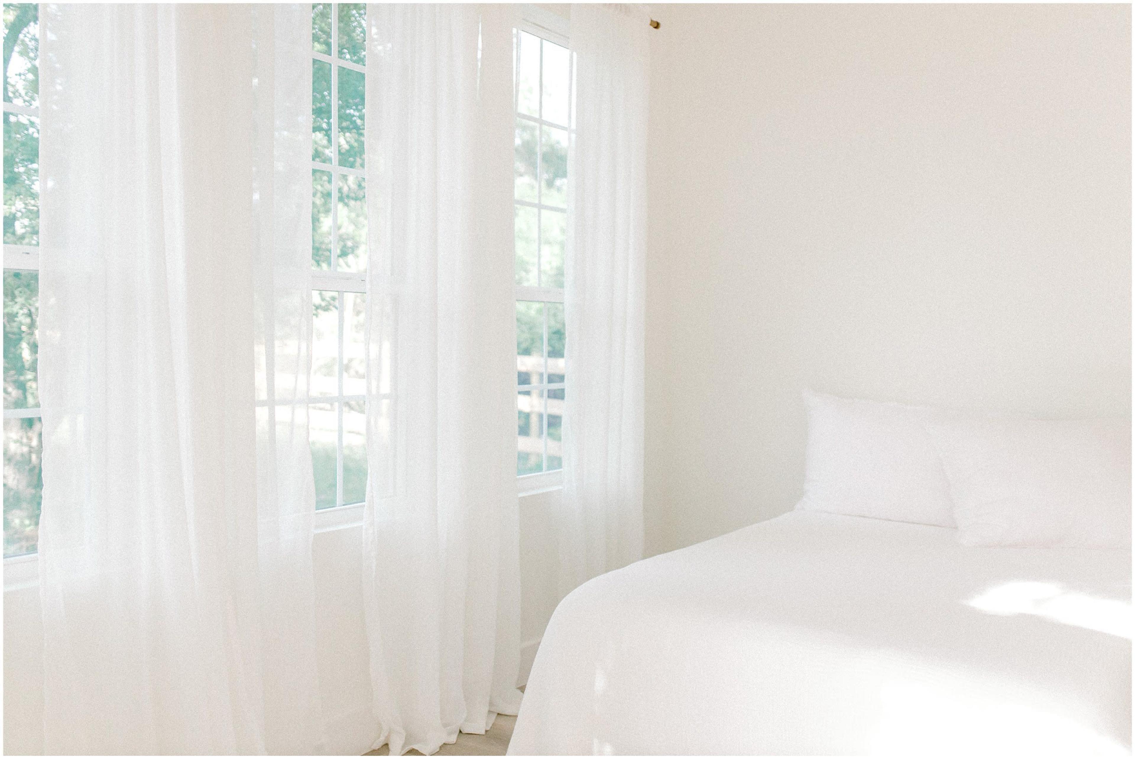 A bright bedroom features a bed with white bedding and large windows adorned with sheer white curtains.