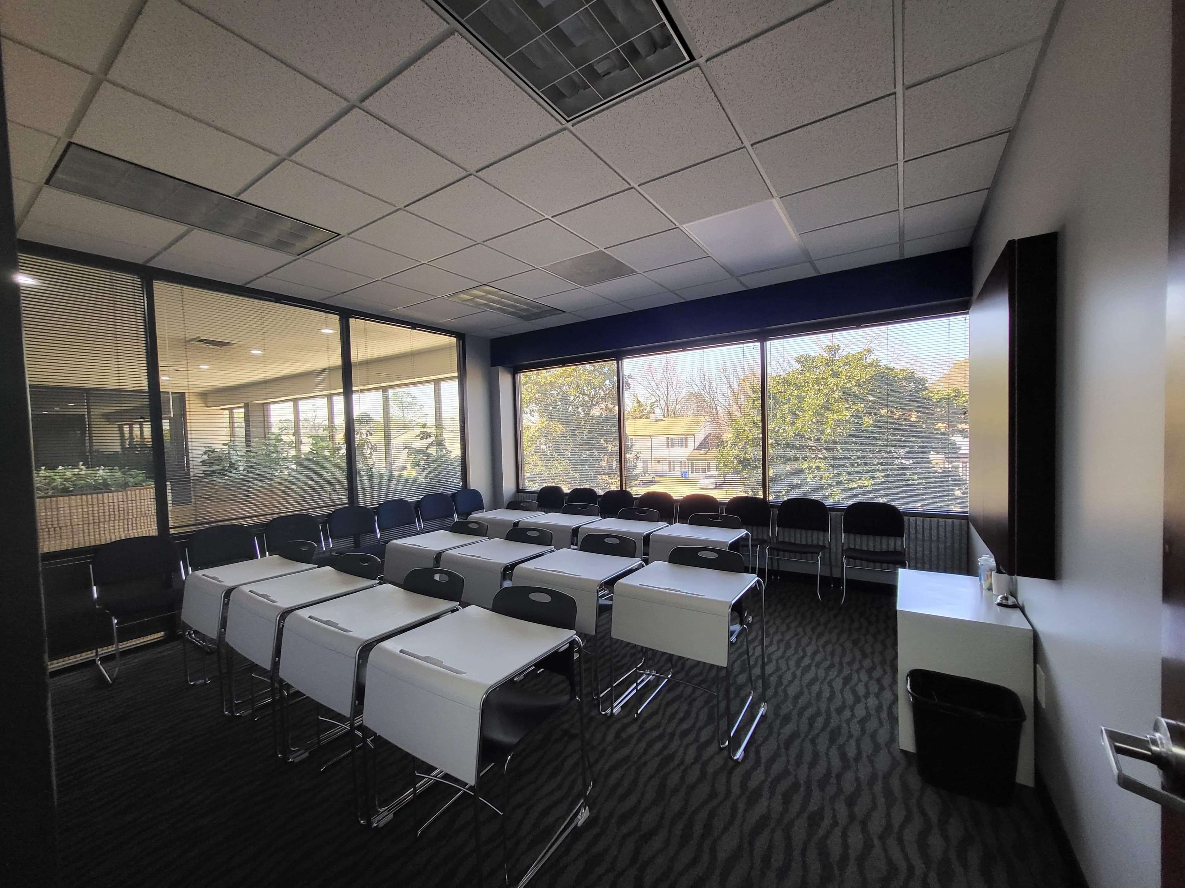 A classroom setup with rows of tables and chairs arranged facing large windows that overlook greenery outside.