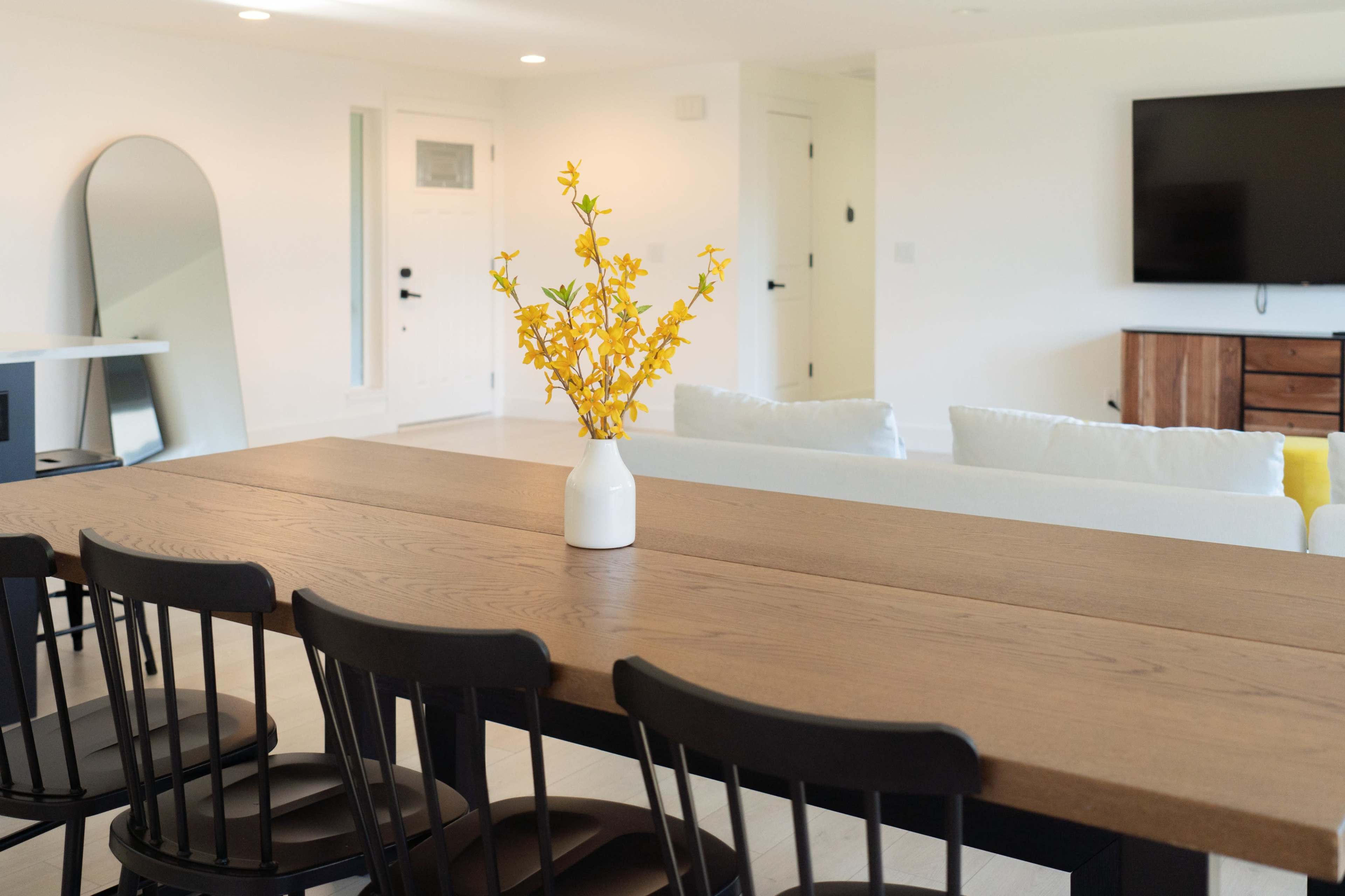A long wooden dining table with black chairs is adorned with a white vase filled with yellow flowers, set in a bright, minimalist living space featuring a large TV and light-colored furnishings.