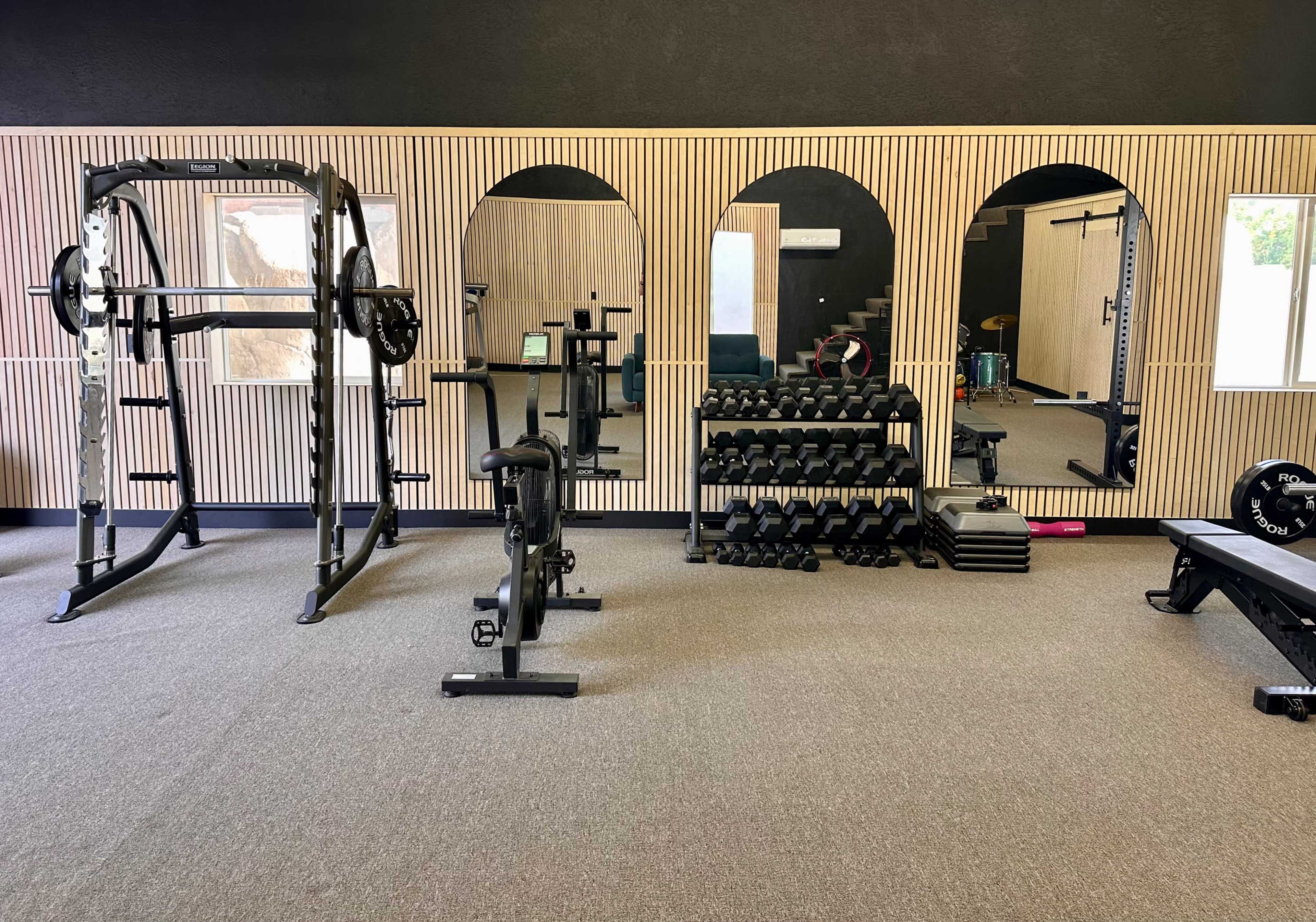 The image shows a gym interior with a squat rack, a rowing machine, a set of dumbbells on a rack, and mirrors along a wooden-paneled wall.