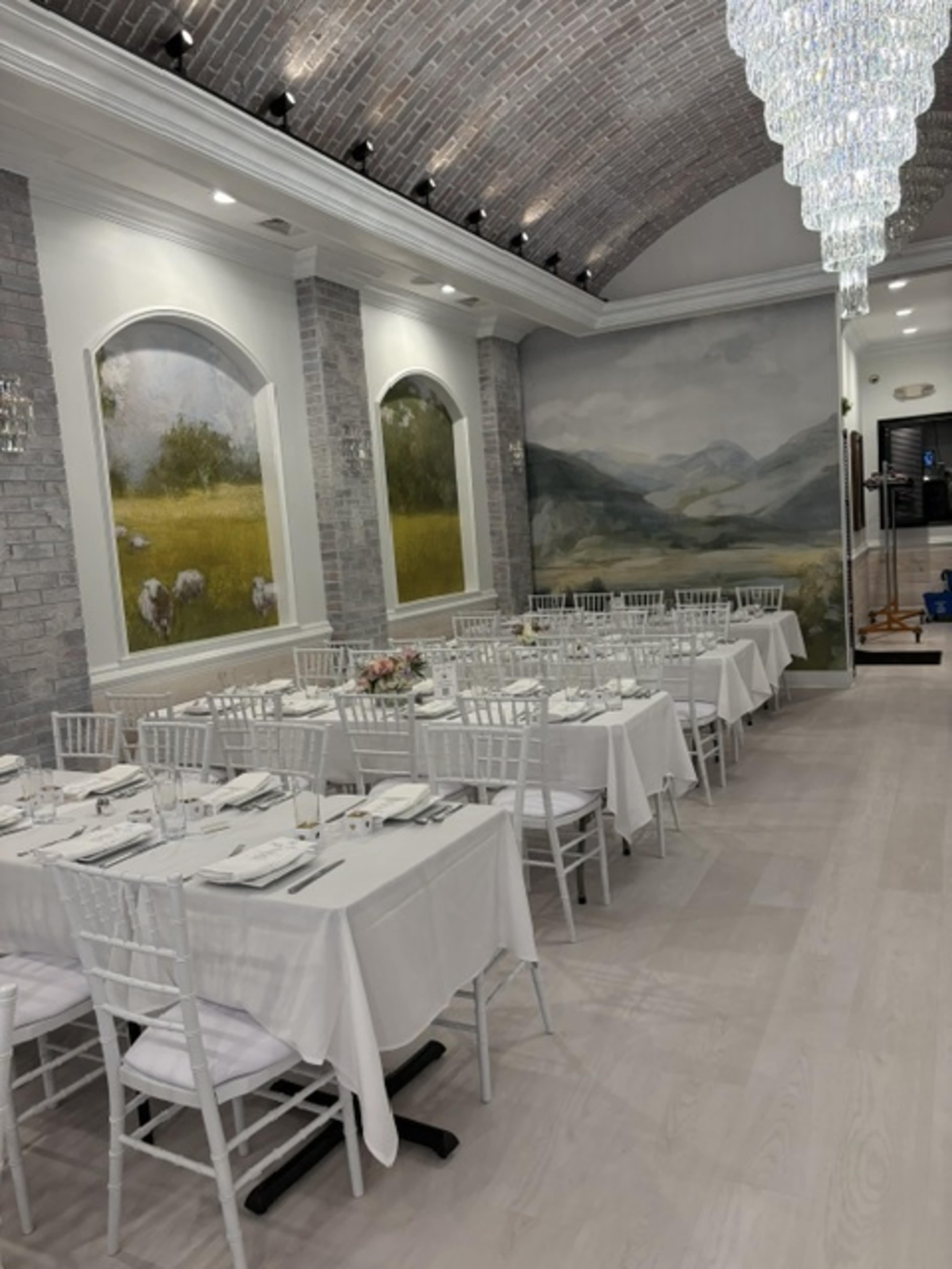 The image shows a elegantly arranged dining room with white tables set for a meal, featuring grey brick walls and decorative light fixtures.