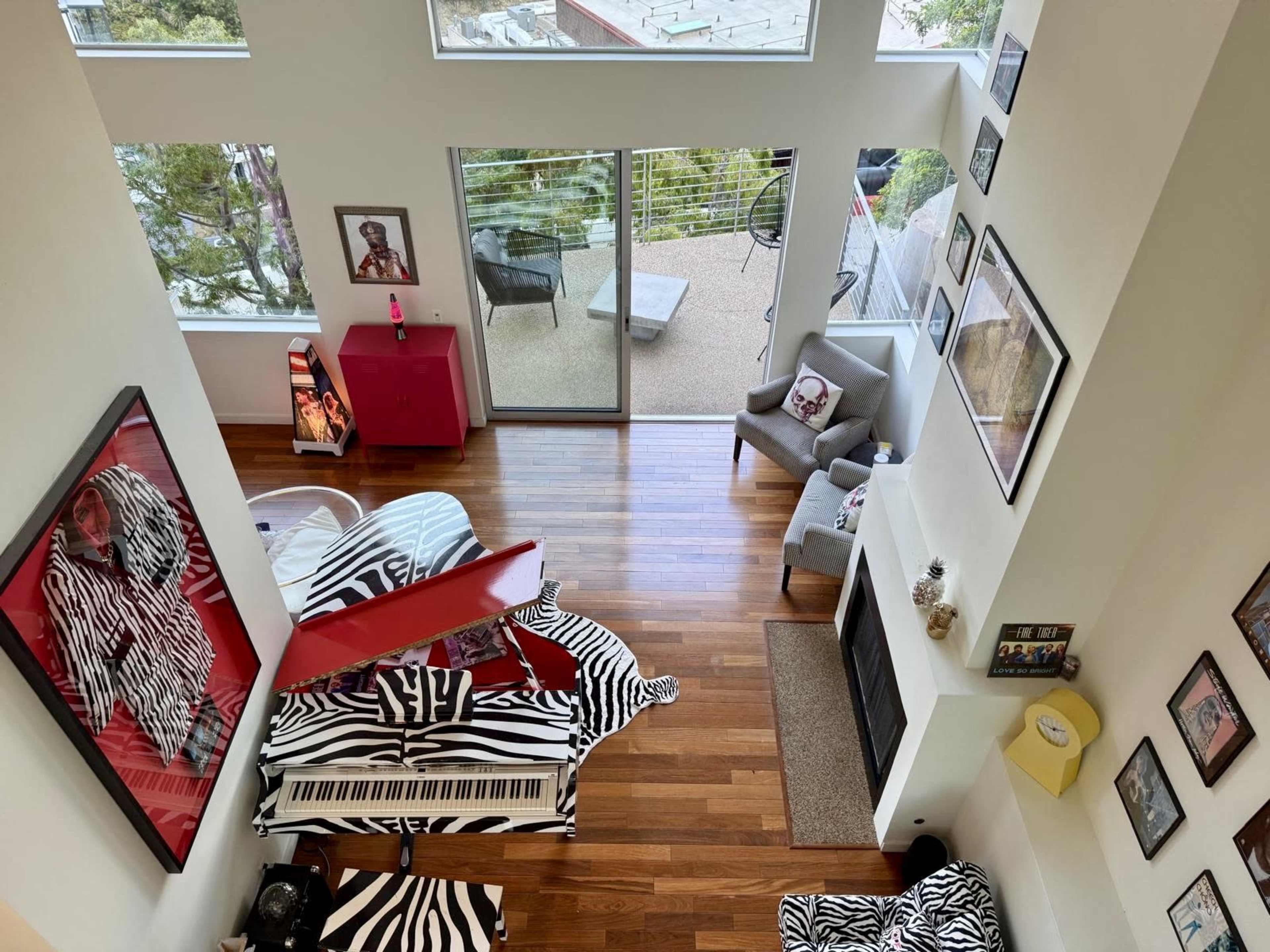 The image shows a modern living space with large windows, wooden flooring, a red piano, and zebra-patterned furniture.