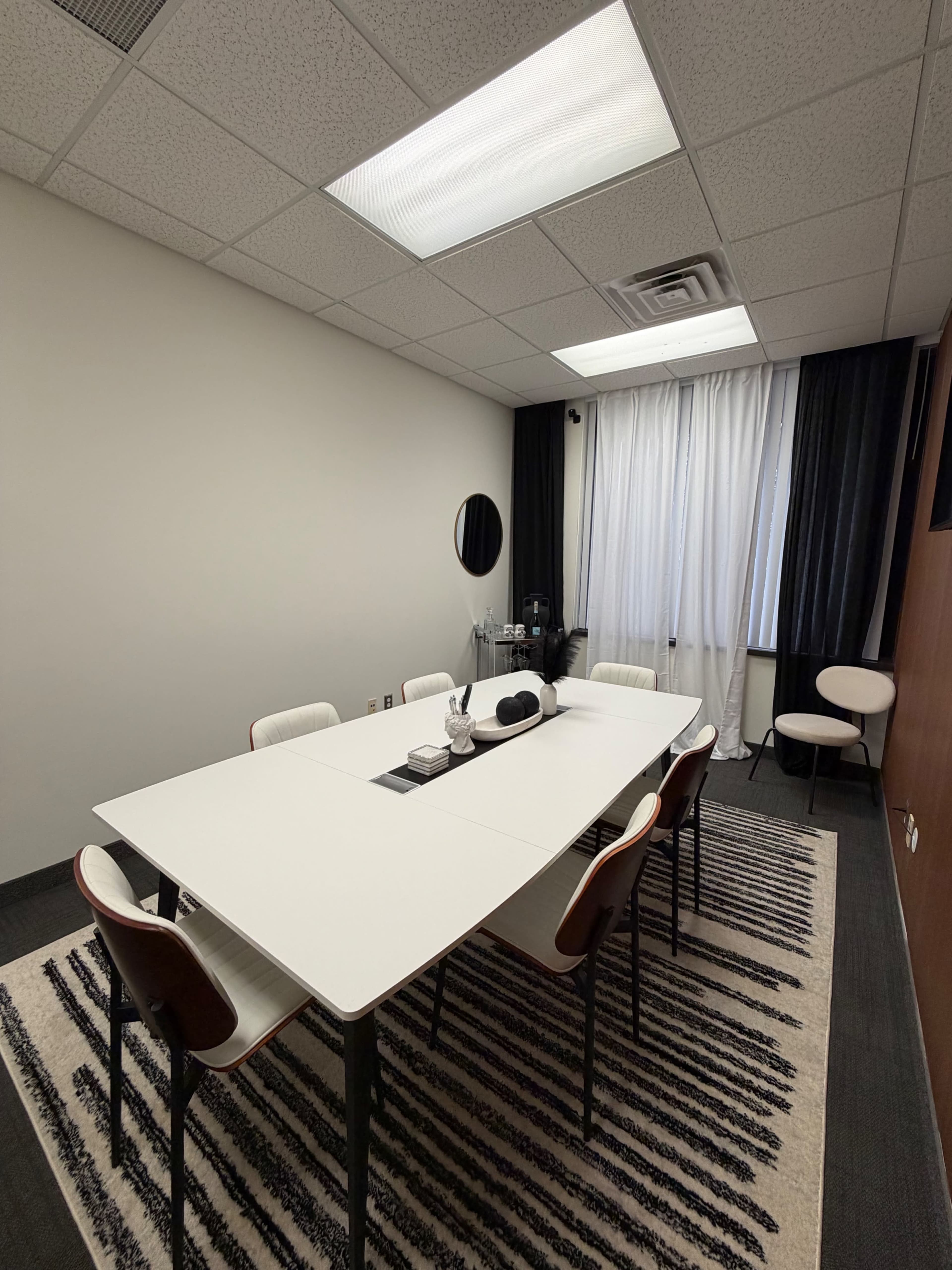 The image shows a modern conference room with a large table surrounded by six chairs and a window covered by white curtains.