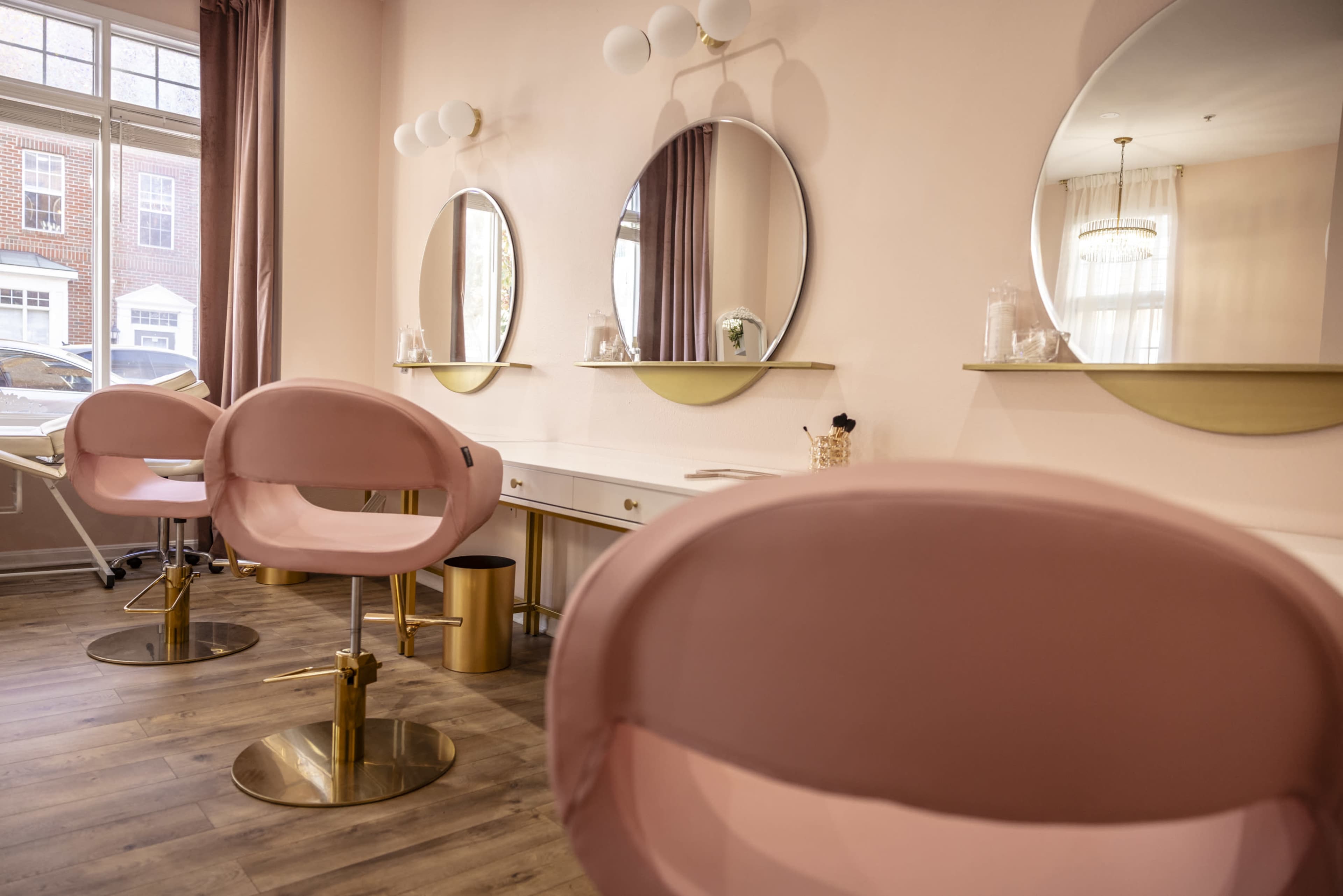 A beauty salon interior with pink chairs and three round mirrors above a white vanity.