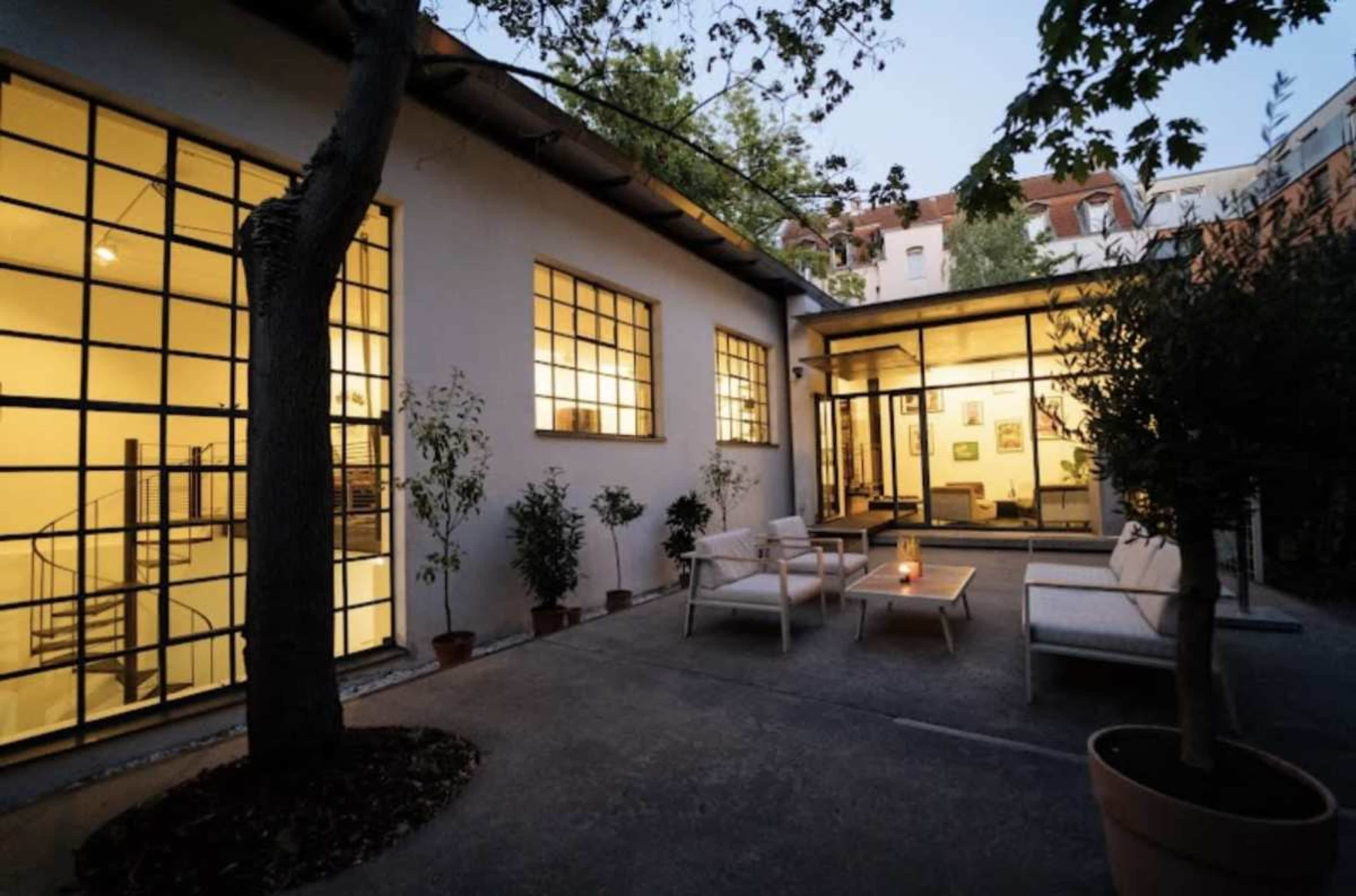 The image shows a modern courtyard of a building with large windows, featuring outdoor seating and potted plants in the evening light.