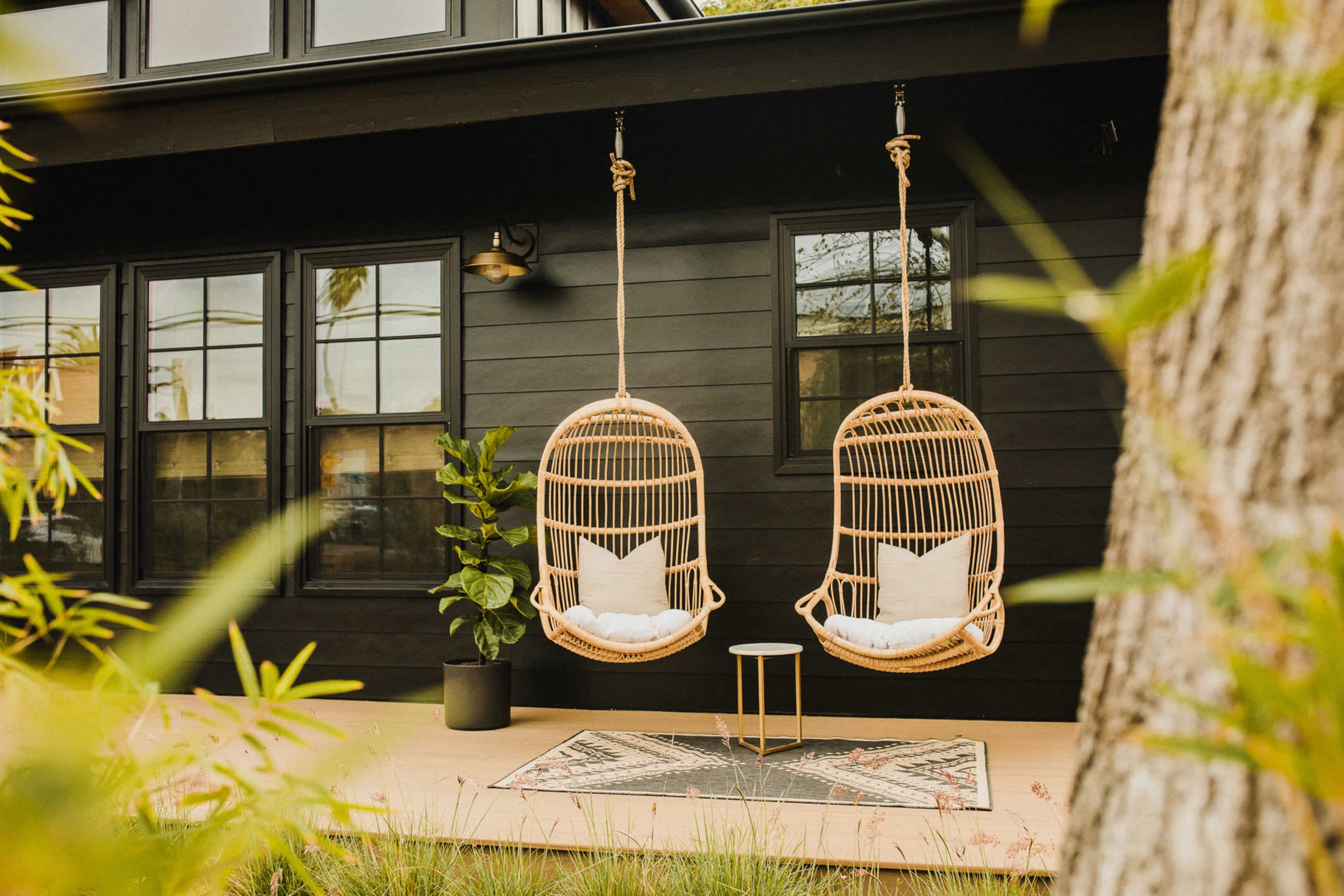 Two suspended rattan chairs with white cushions hang in front of a black house, accompanied by a small table and a decorative rug.