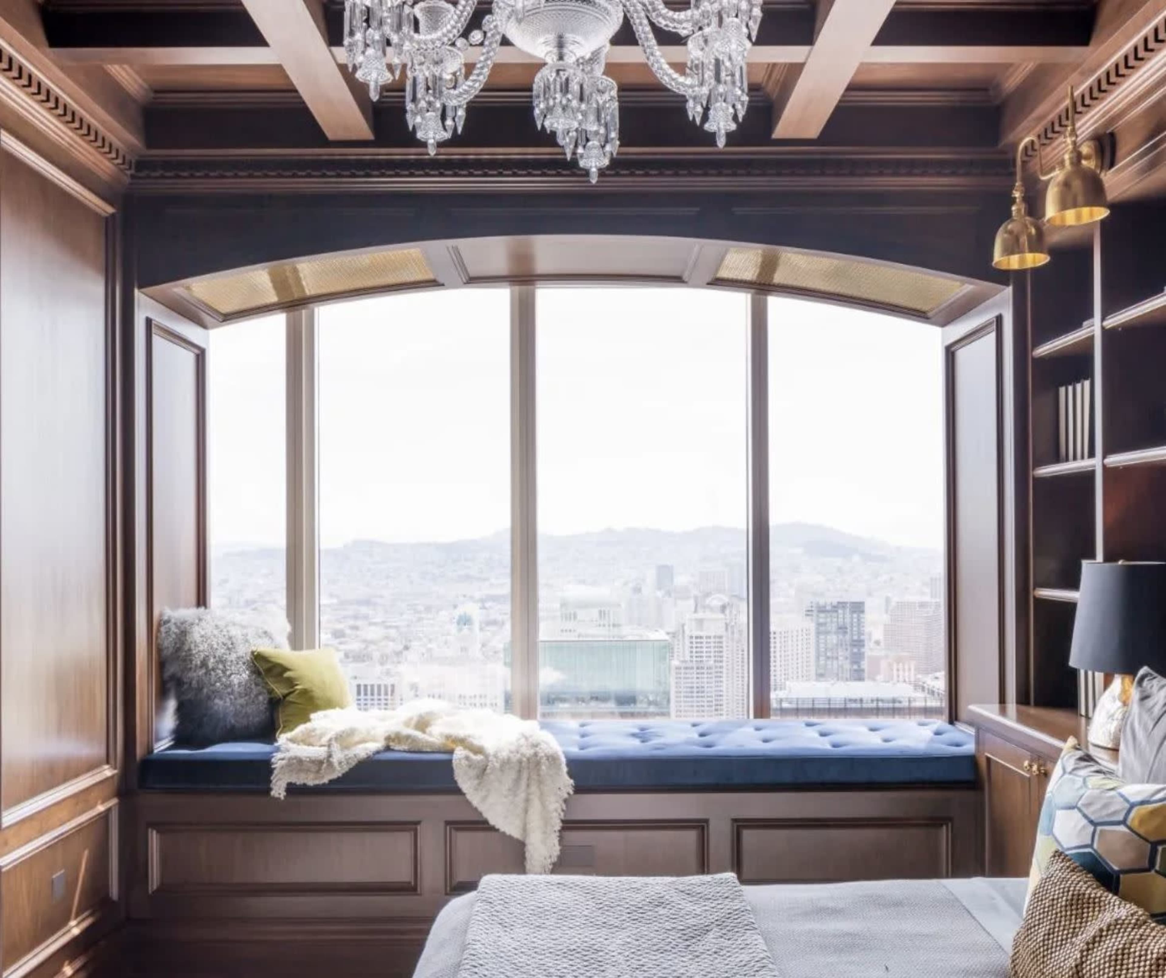 A bedroom with a large window seat overlooking a city skyline, featuring wooden paneling and a chandelier.