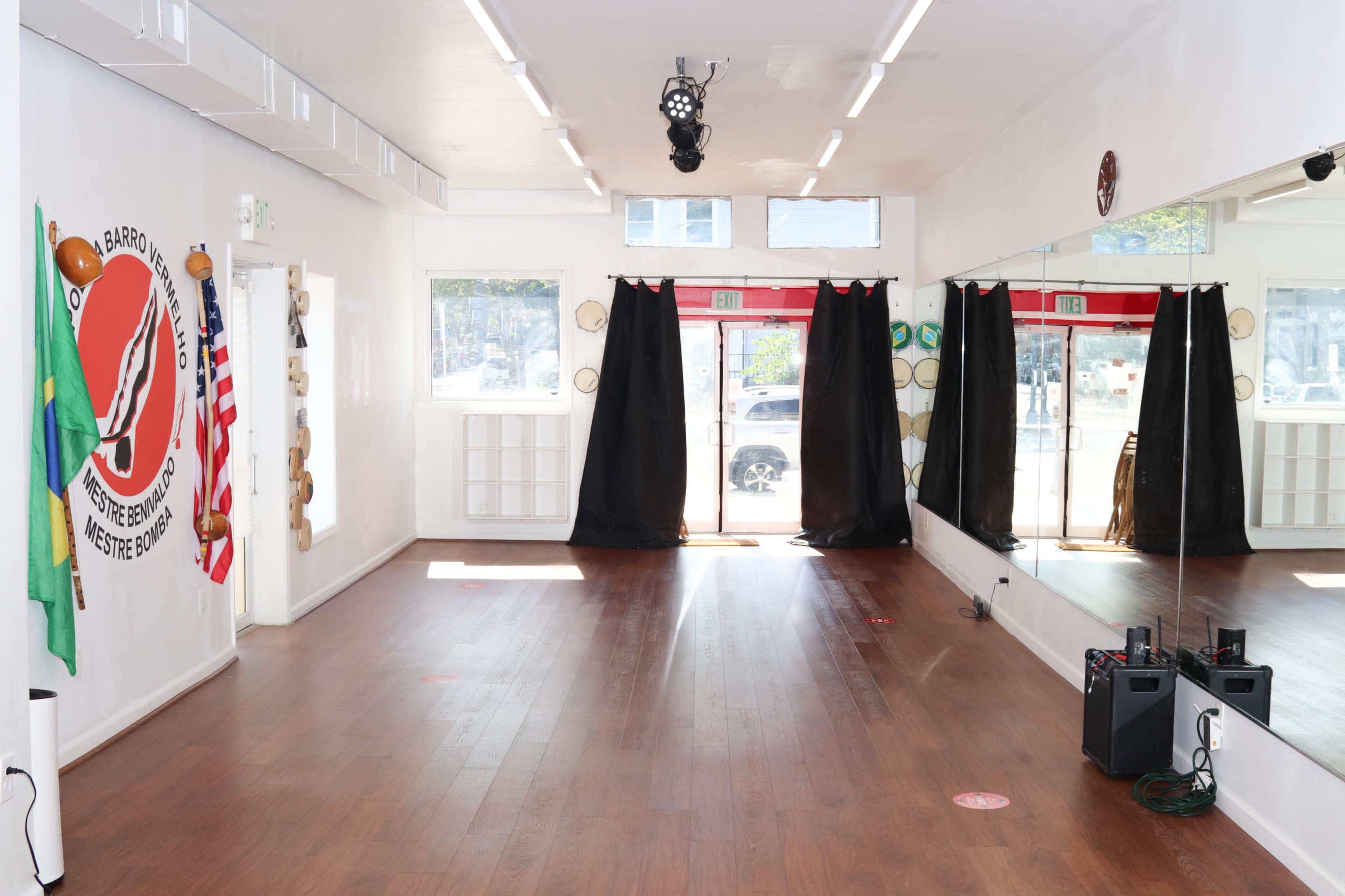 A spacious dance studio with wooden flooring, mirrored walls, and black curtains, adorned with flags and a logo on one wall.