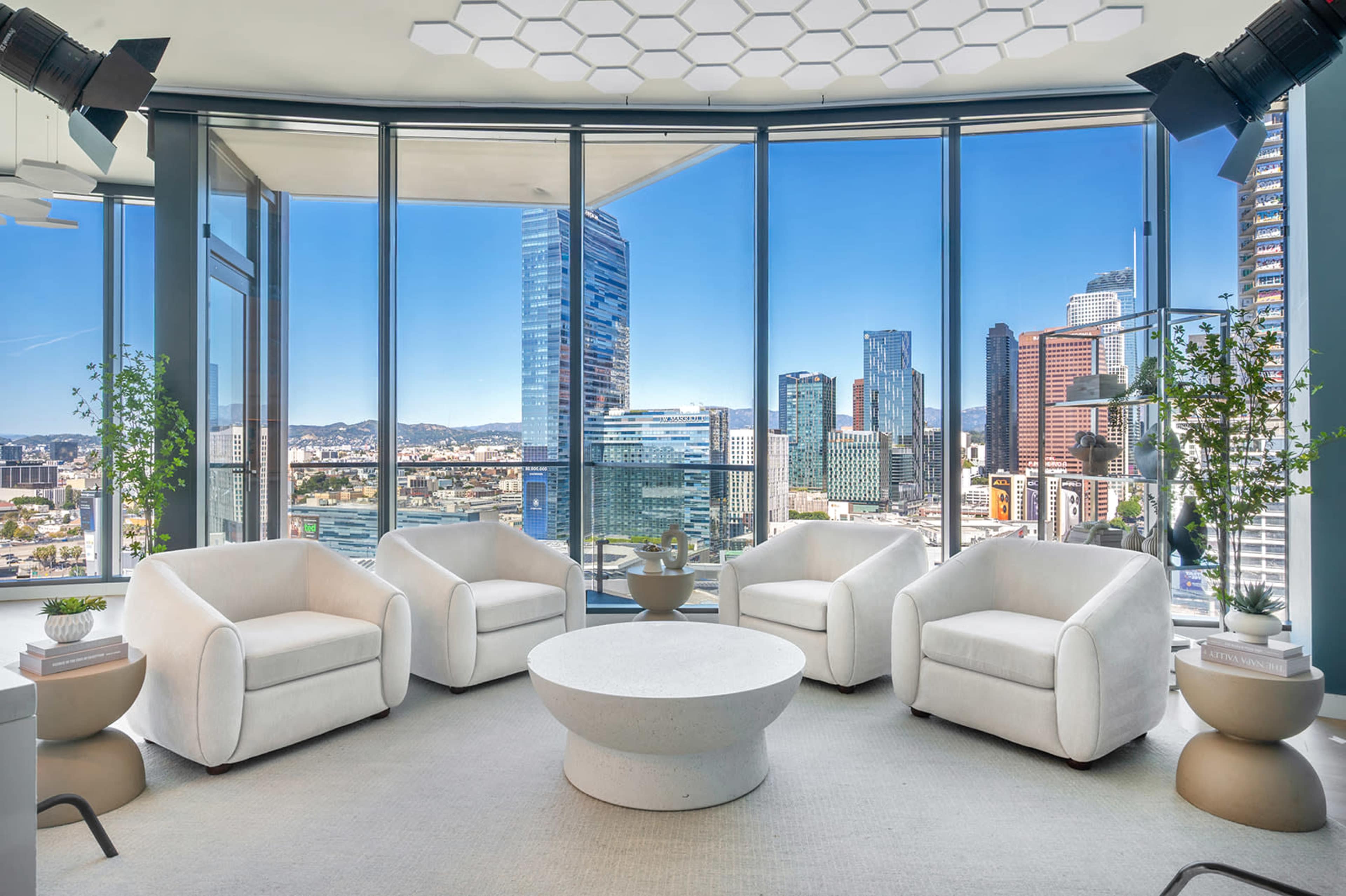A modern lounge area features four white chairs and a round coffee table, with large windows offering a panoramic view of a city skyline.