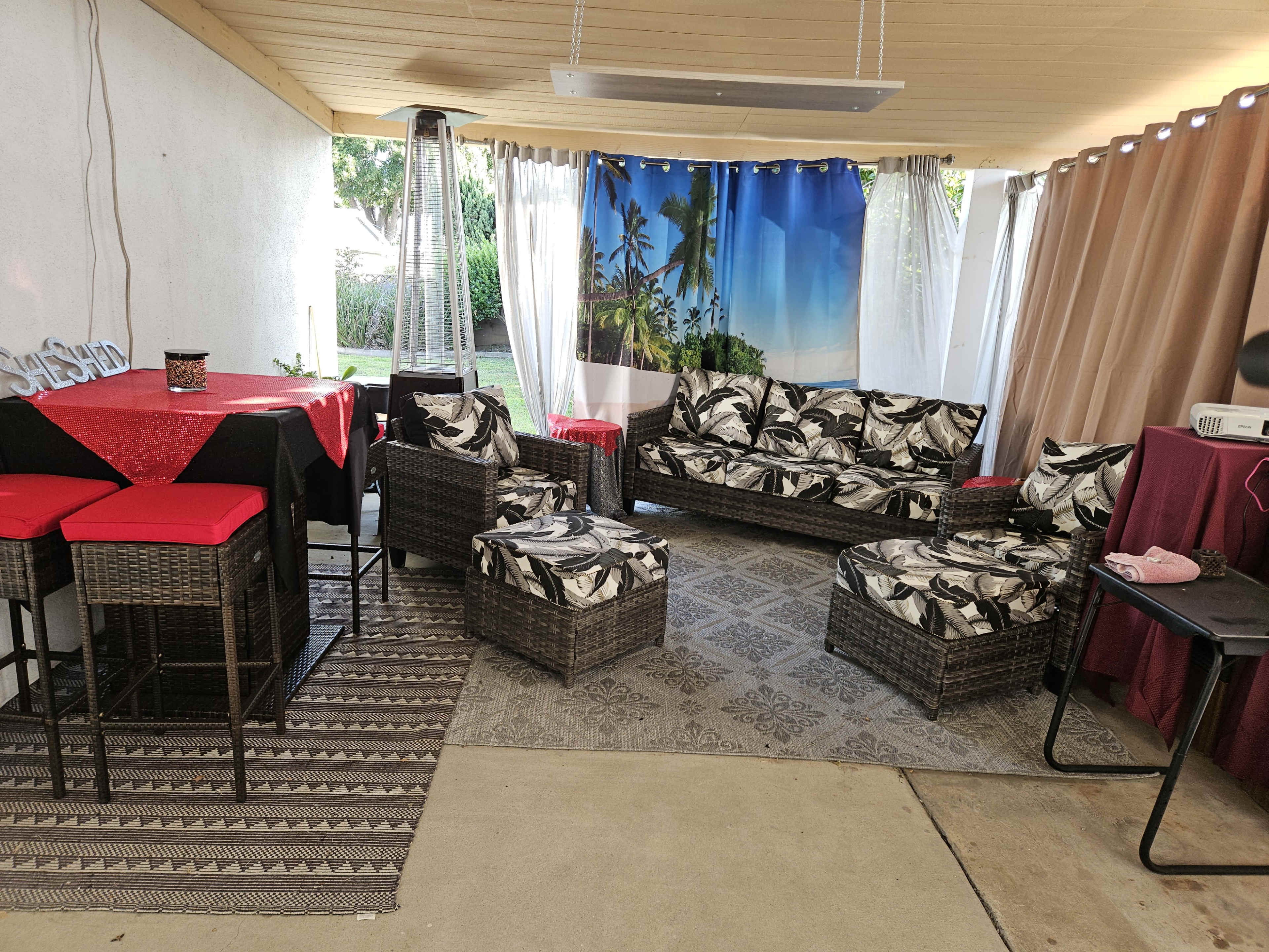 A covered outdoor seating area with wicker furniture, a red table, and a backdrop featuring palm trees and a blue sky.