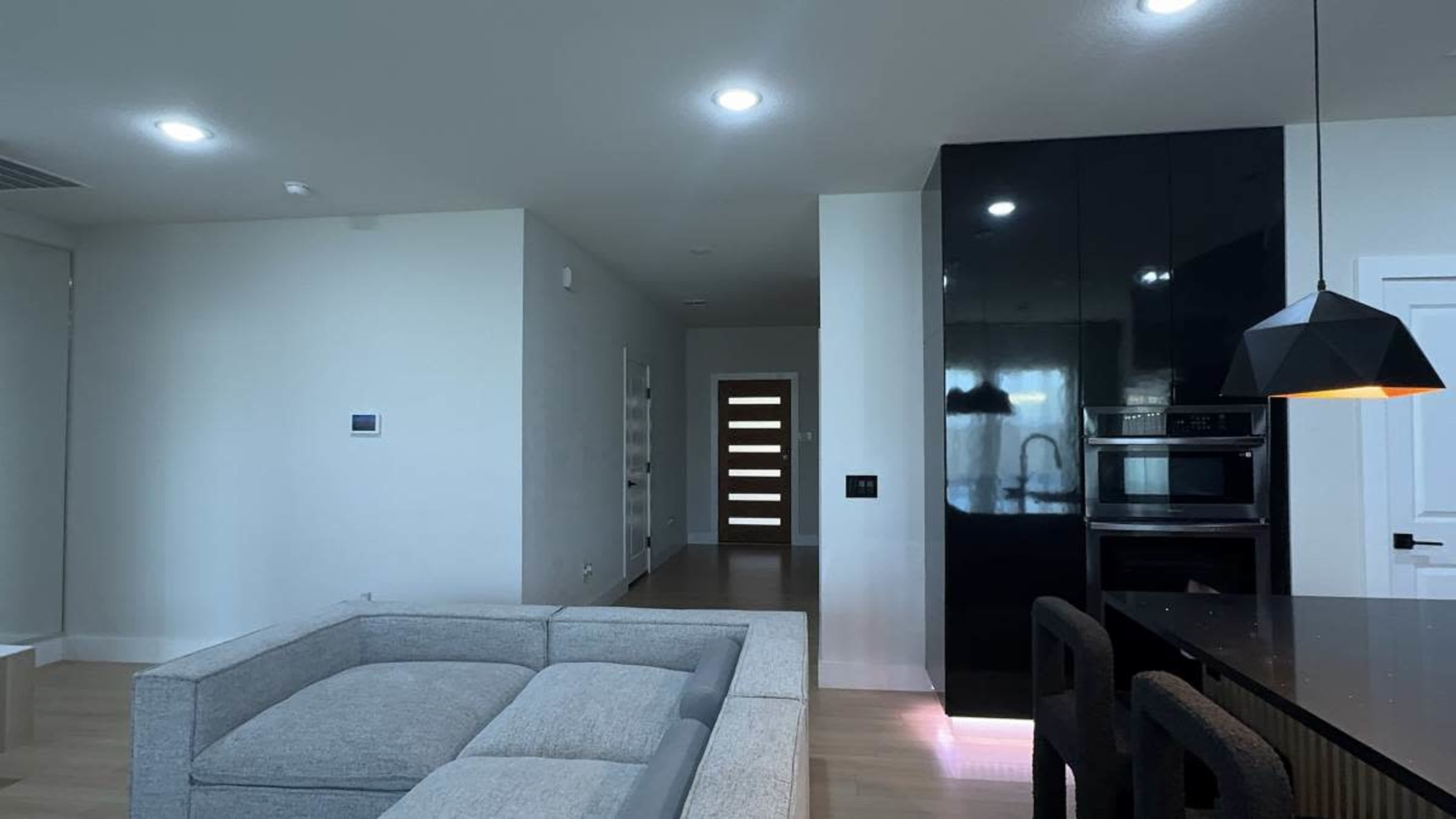 The image shows a modern, open-concept living space featuring a light-colored sofa, a sleek kitchen with black cabinetry, and a view of a hallway with a wooden door.