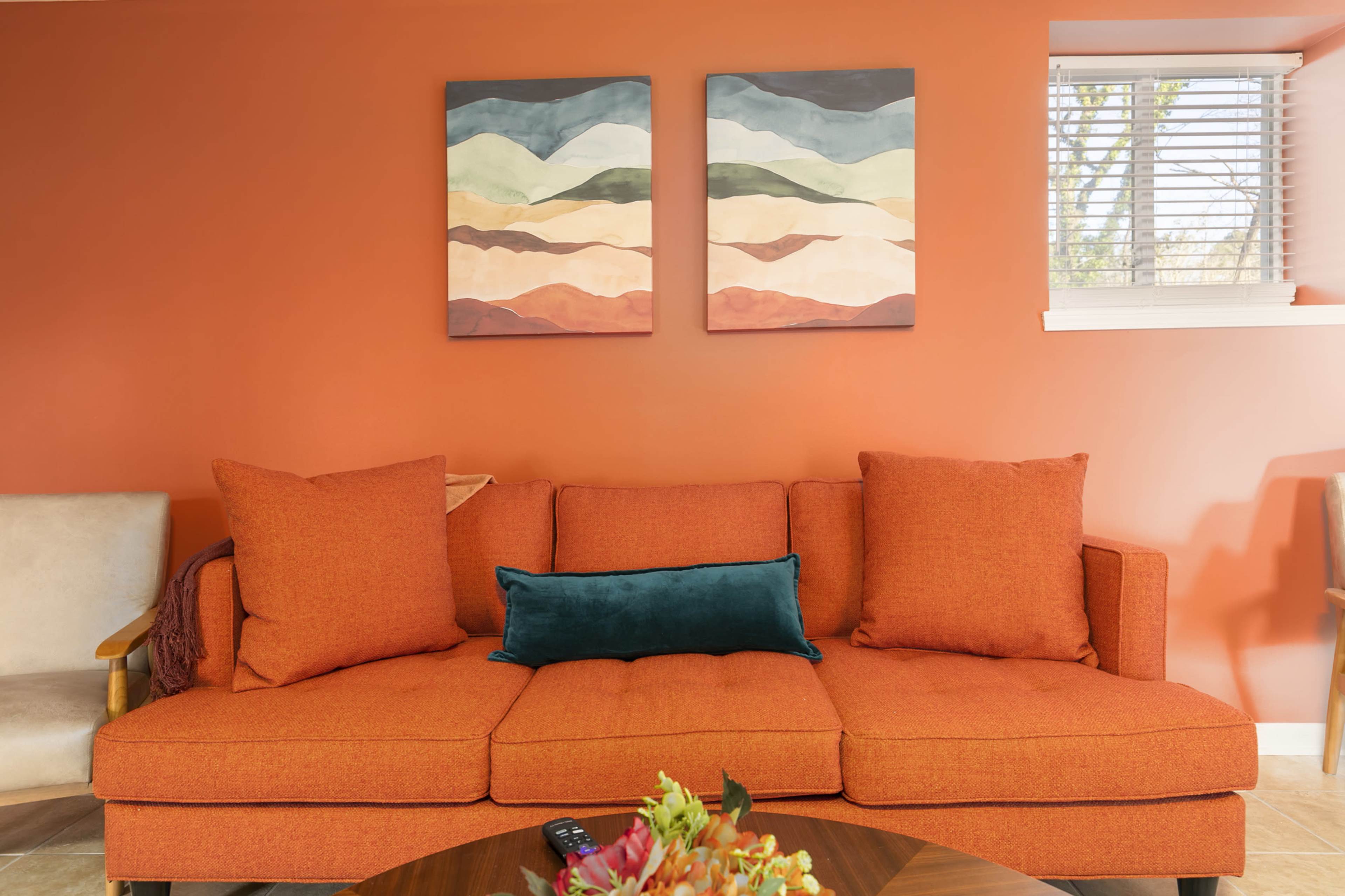 A rust-colored sofa with large cushions is positioned against a bright orange wall, adorned with two abstract paintings, while a round coffee table with a floral arrangement sits in front.