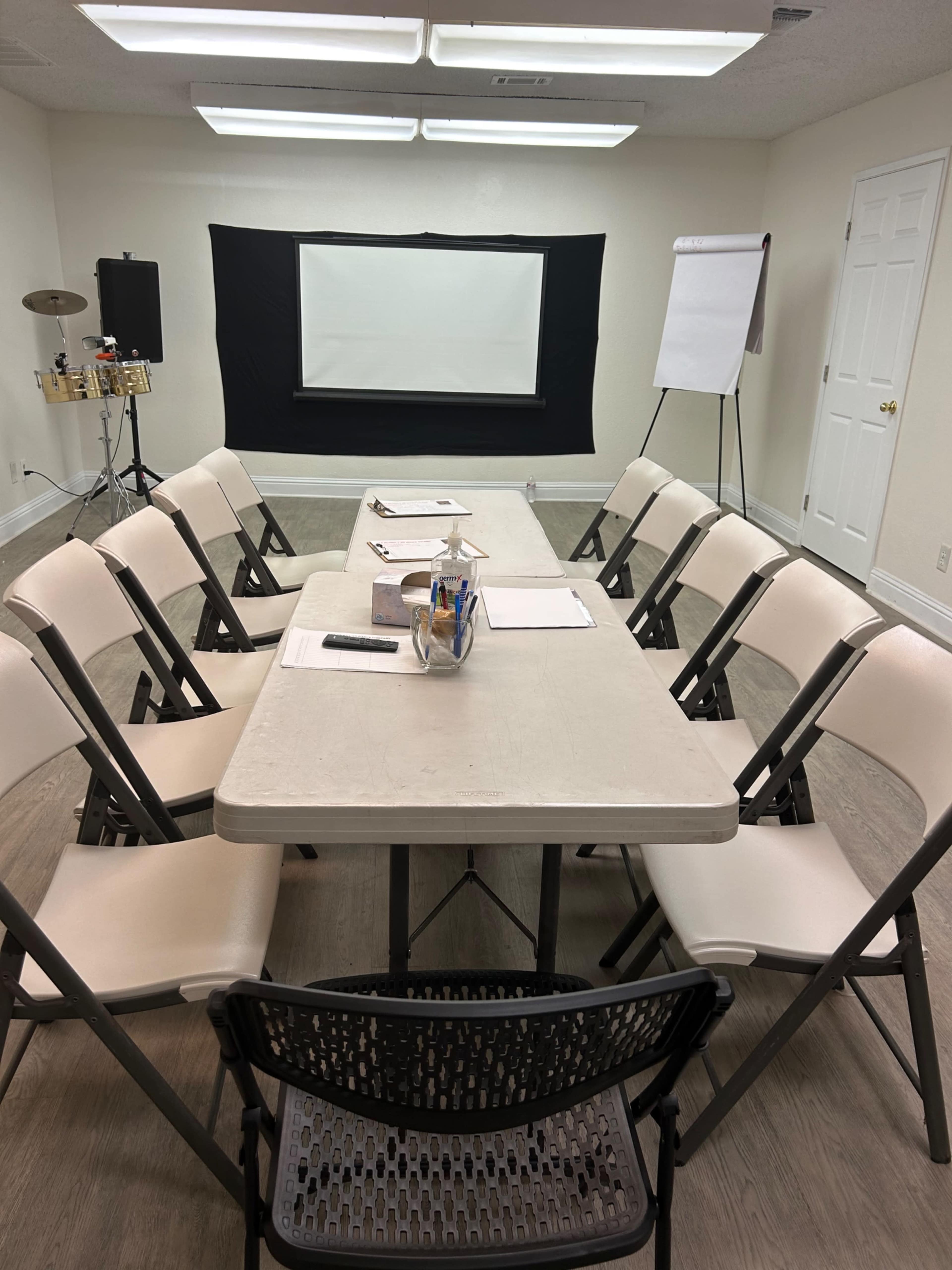 A rectangular table surrounded by folding chairs is set up in a room with a projector screen and a whiteboard for a meeting.
