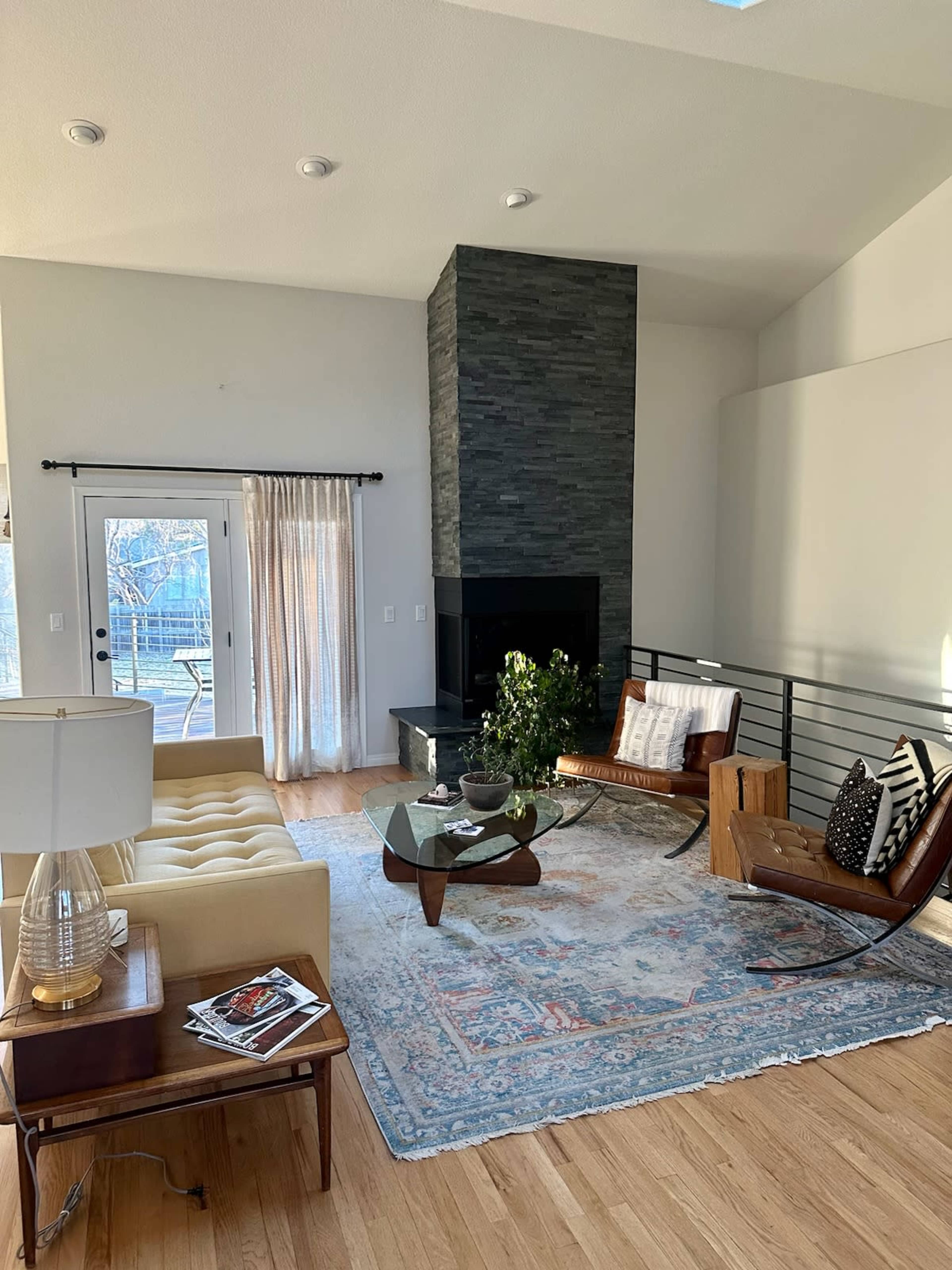 A modern living room features a yellow sofa, a glass coffee table, a stone fireplace, and large windows with sheer curtains.