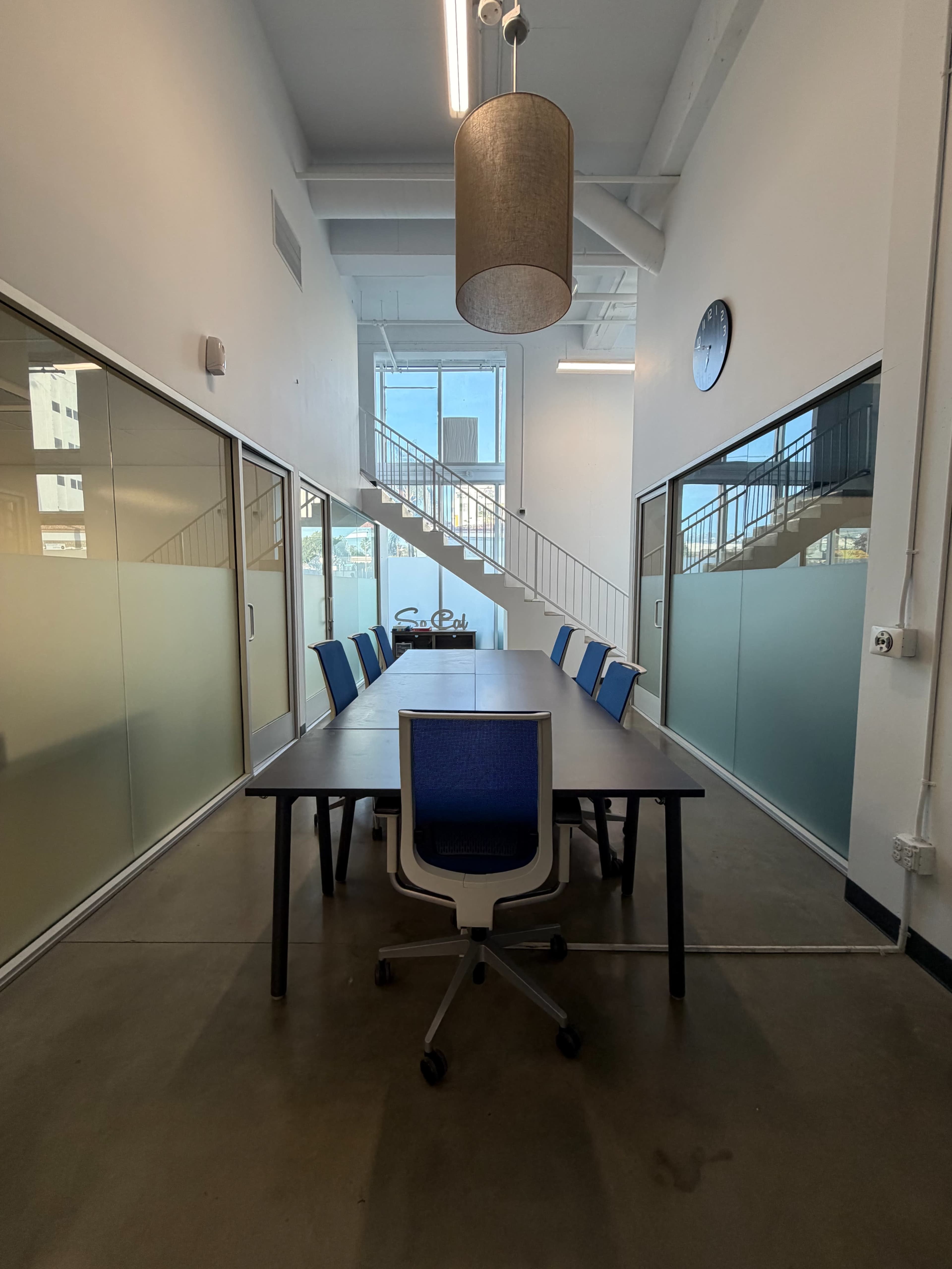 A modern conference room features a long table with blue chairs, large windows, and a staircase in the background.