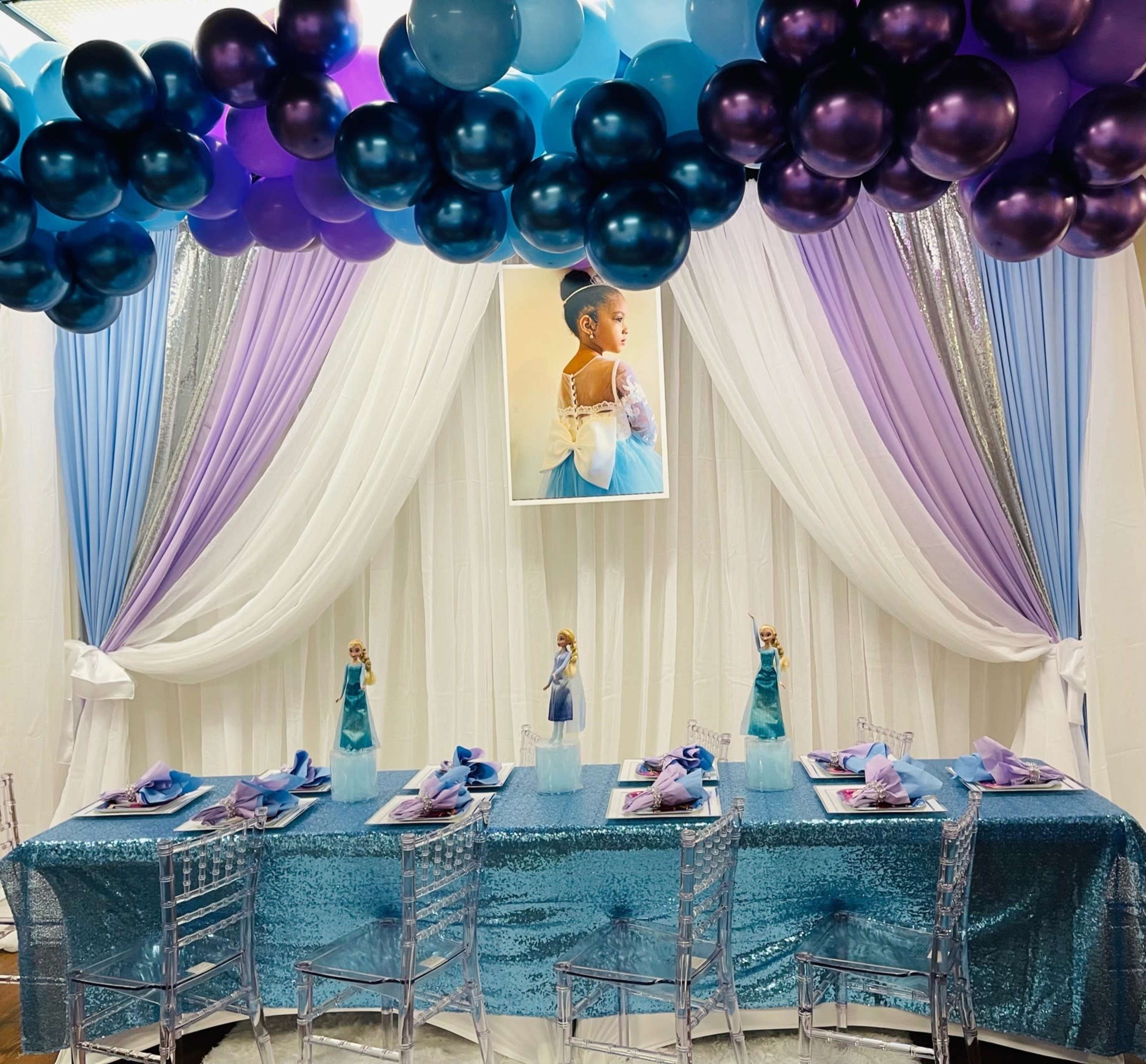 A party table is set with a shimmering blue tablecloth, adorned with place settings and decorative figures, under a balloon arch in purple and blue hues.