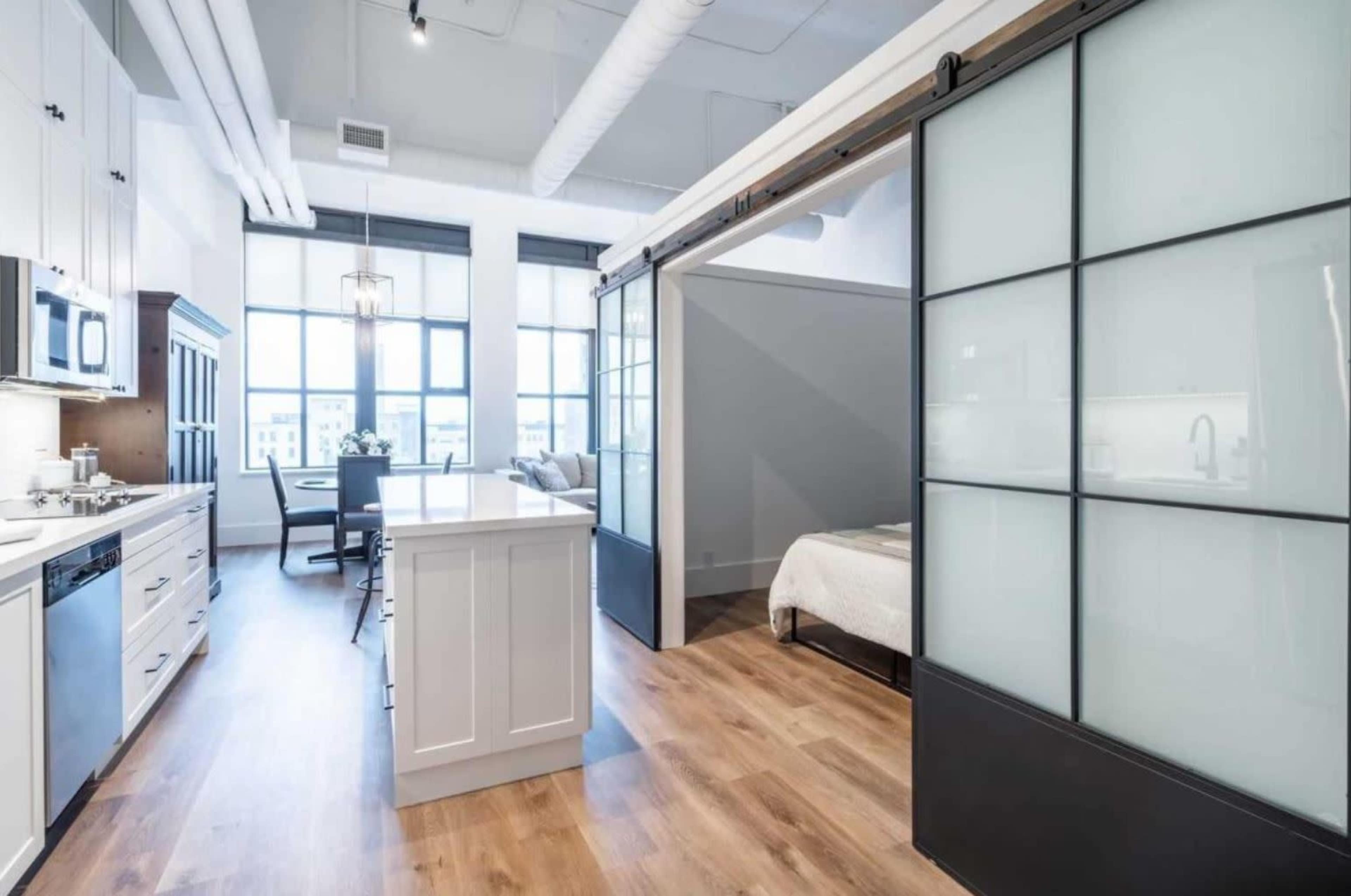 A modern open-space apartment featuring a kitchen island, sliding glass doors separating the living area from a bedroom, and large windows allowing natural light to enter.