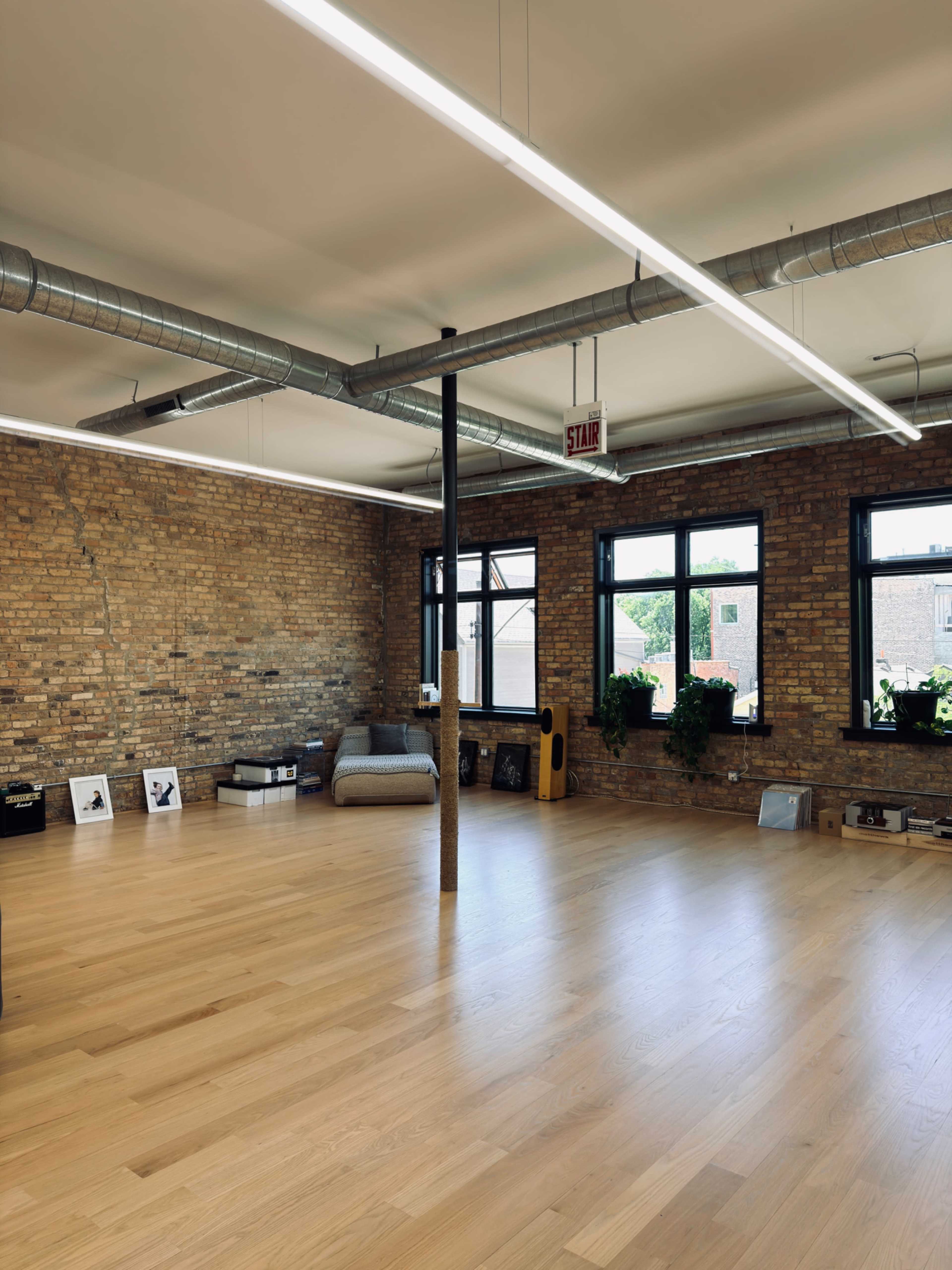 The image shows a spacious room with hardwood flooring, exposed brick walls, large windows, and minimal furnishings.