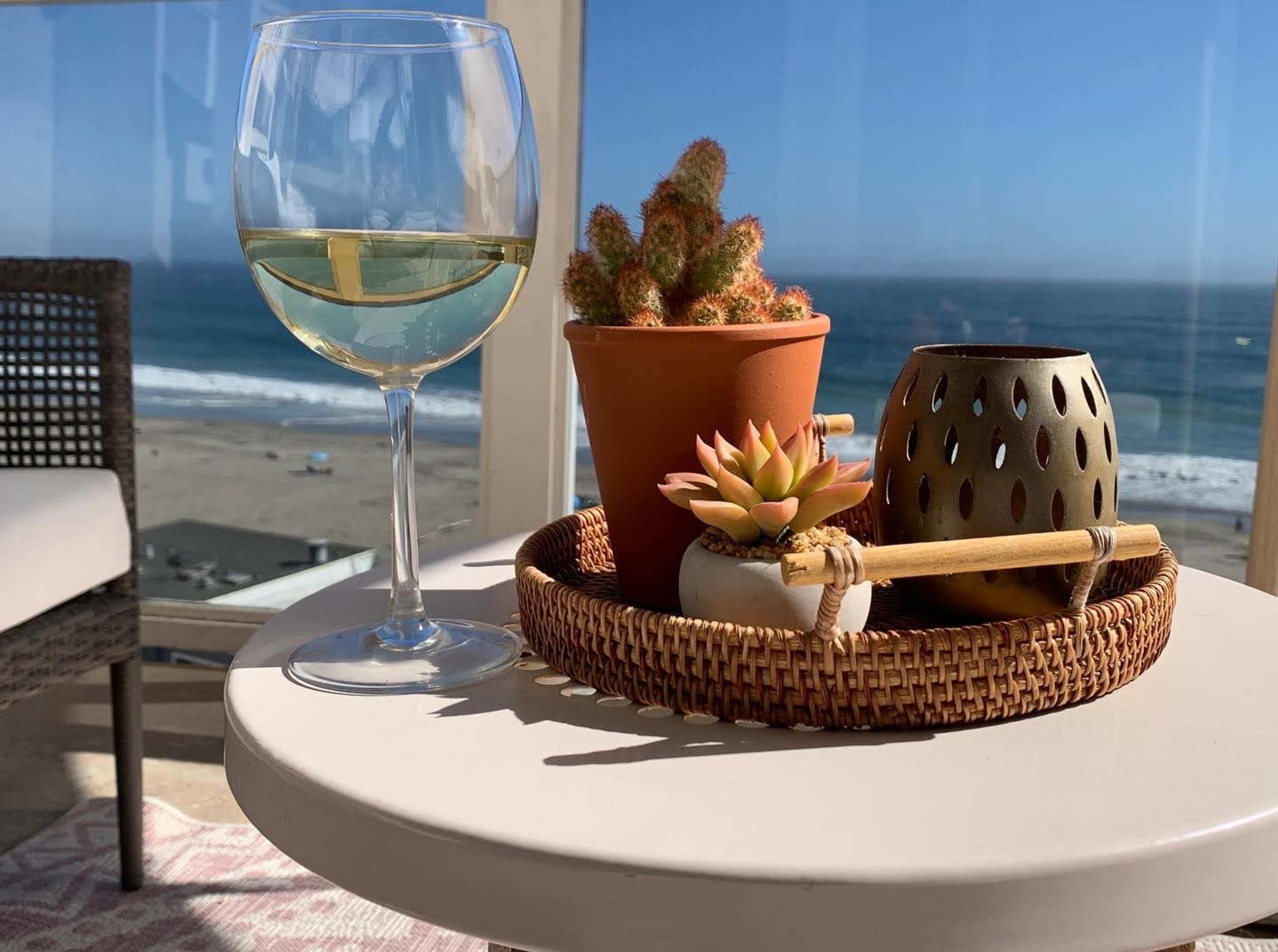 A glass of white wine rests on a round table alongside a succulent plant in a terracotta pot and a decorative candle holder, with a view of the ocean in the background.