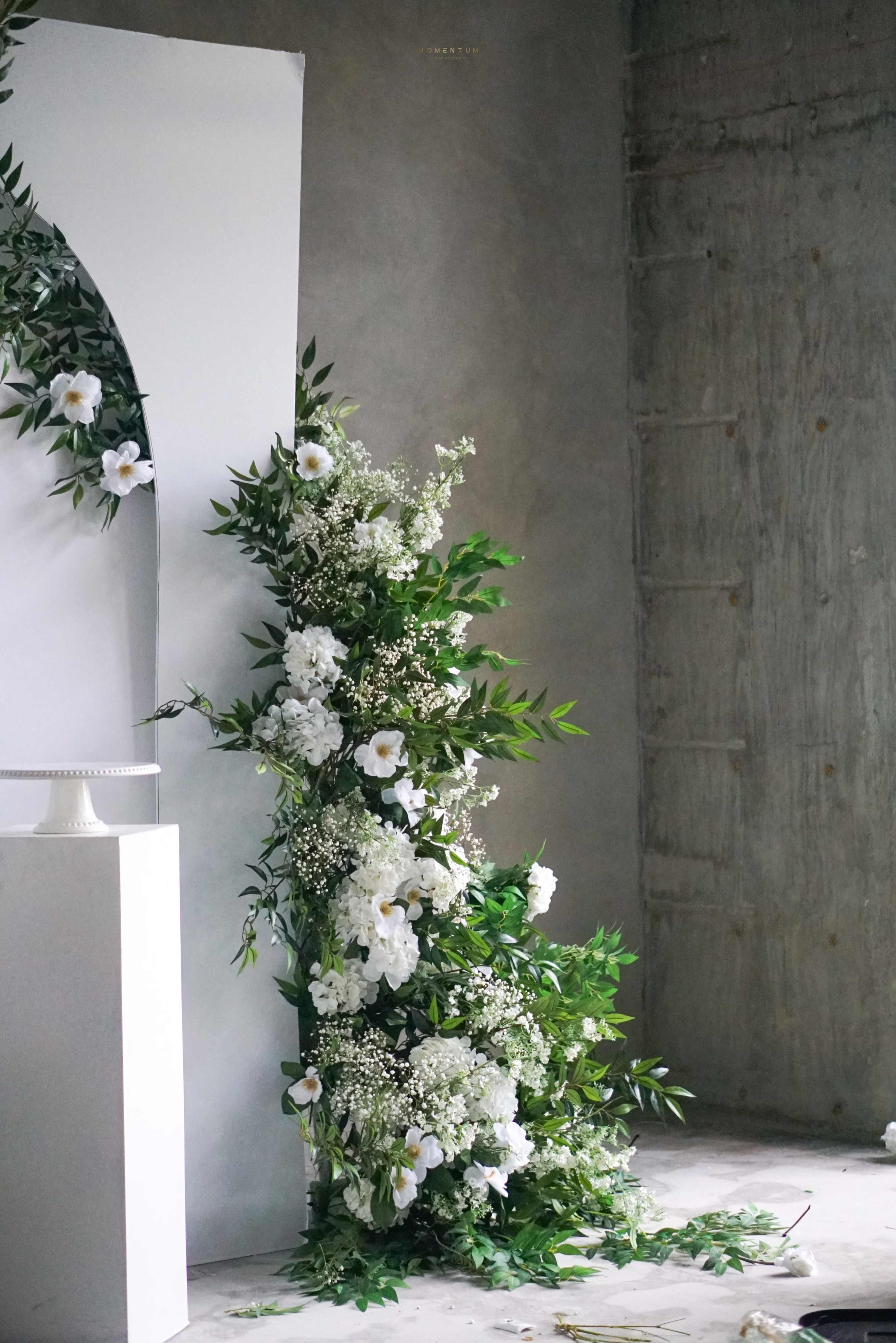 A floral arrangement featuring white flowers and greenery is positioned near a white arch against a concrete wall.