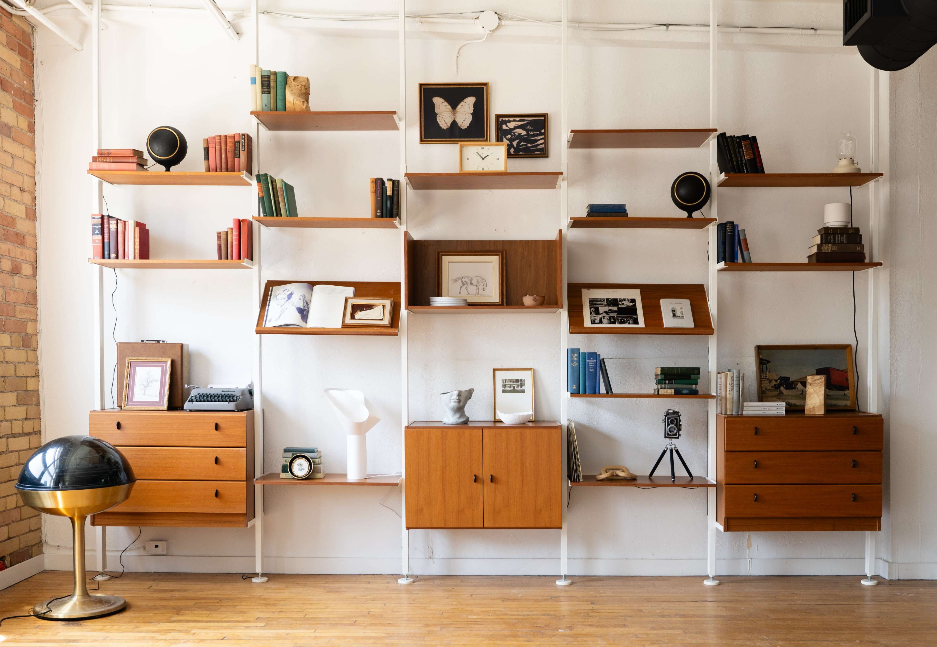 The image shows a modern, minimalist wooden shelving unit with various books, decorative items, and a globe in a bright room.