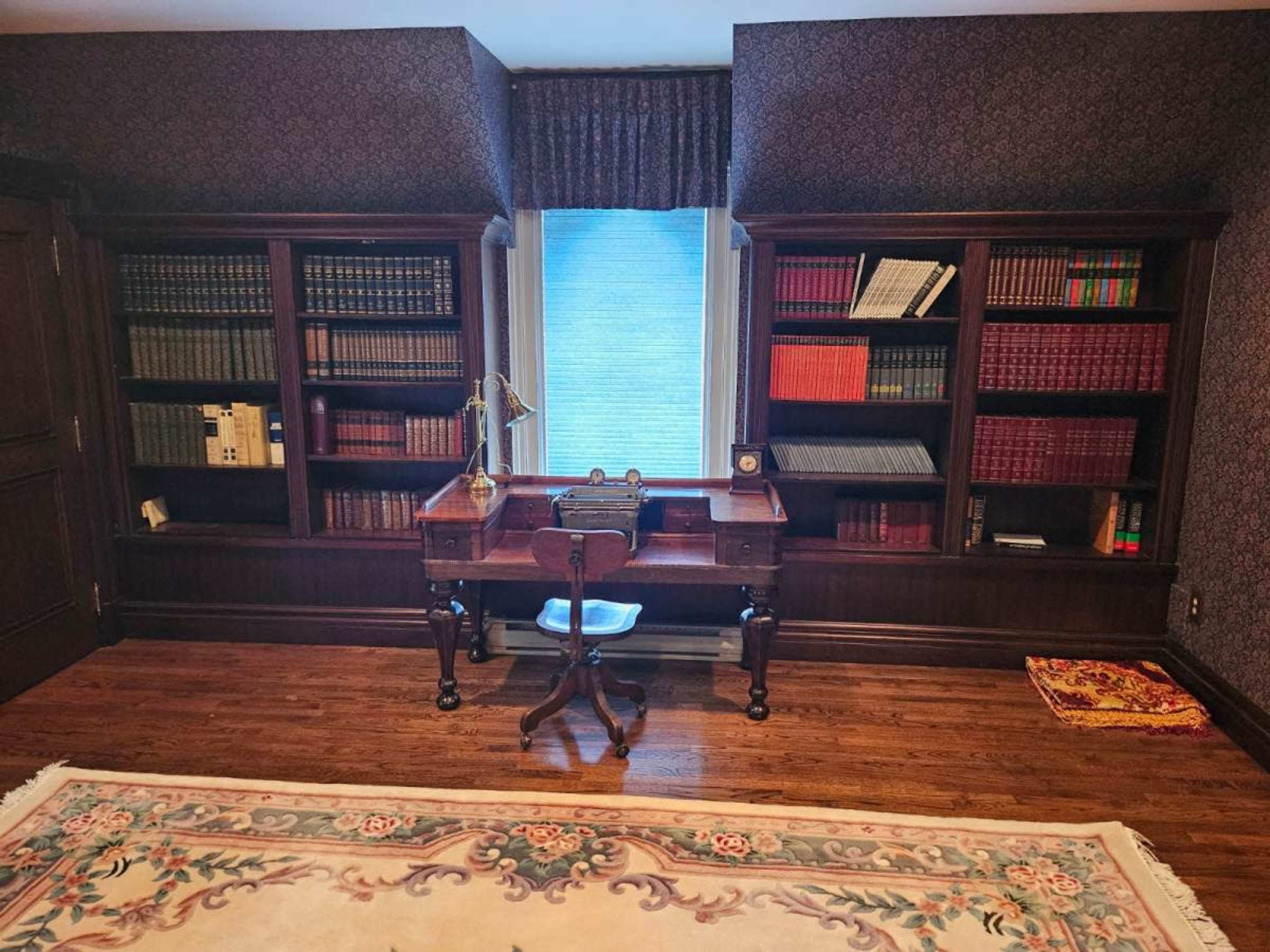 A wooden desk with a chair in front of a window, surrounded by dark wood bookshelves filled with books and decorative items in a room with patterned wallpaper and a patterned rug.