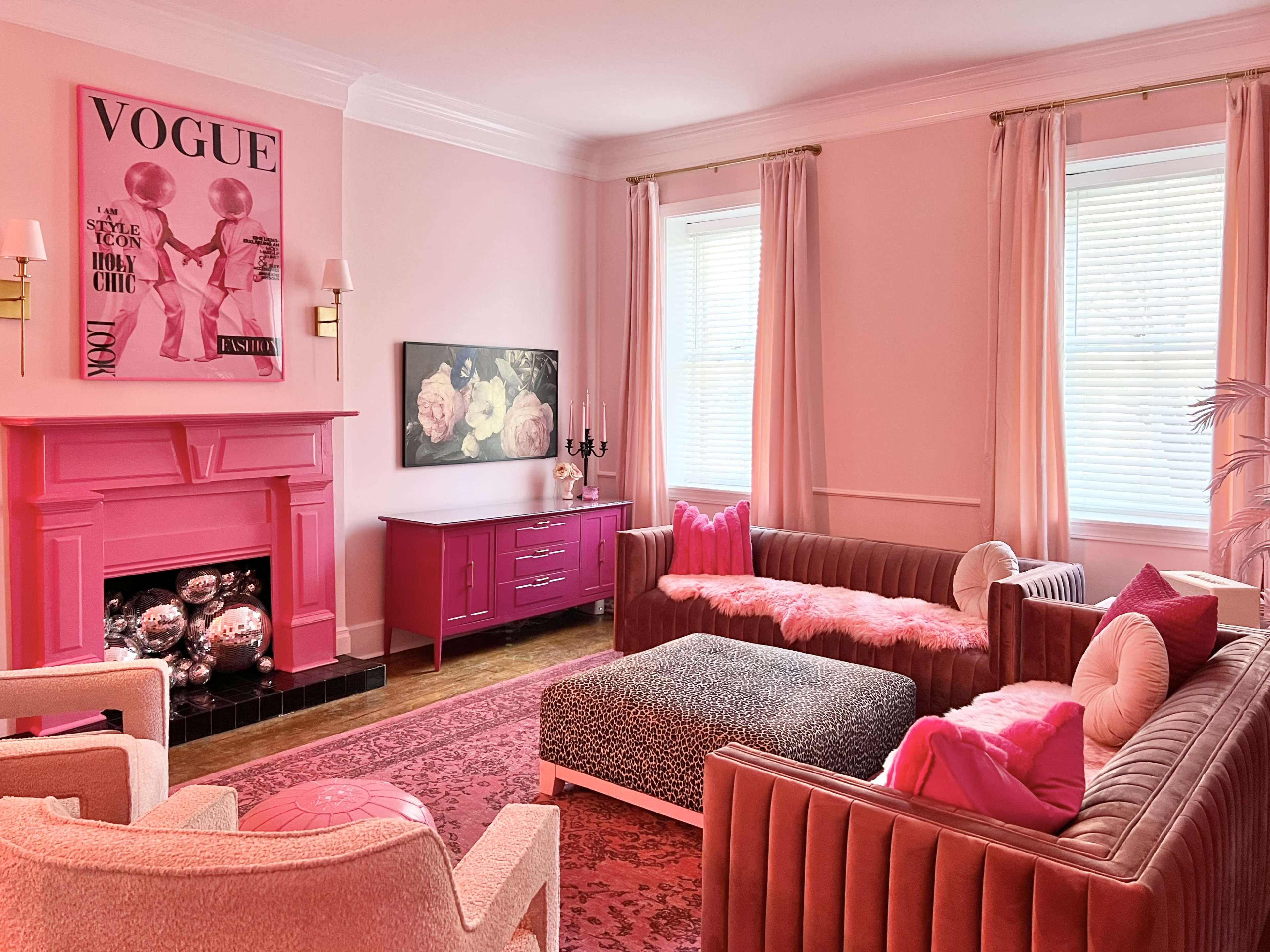 The room features pink walls, a pink fireplace, and furniture that includes two sofas, a patterned ottoman, and a decorative cabinet, complemented by artwork and floral decor.