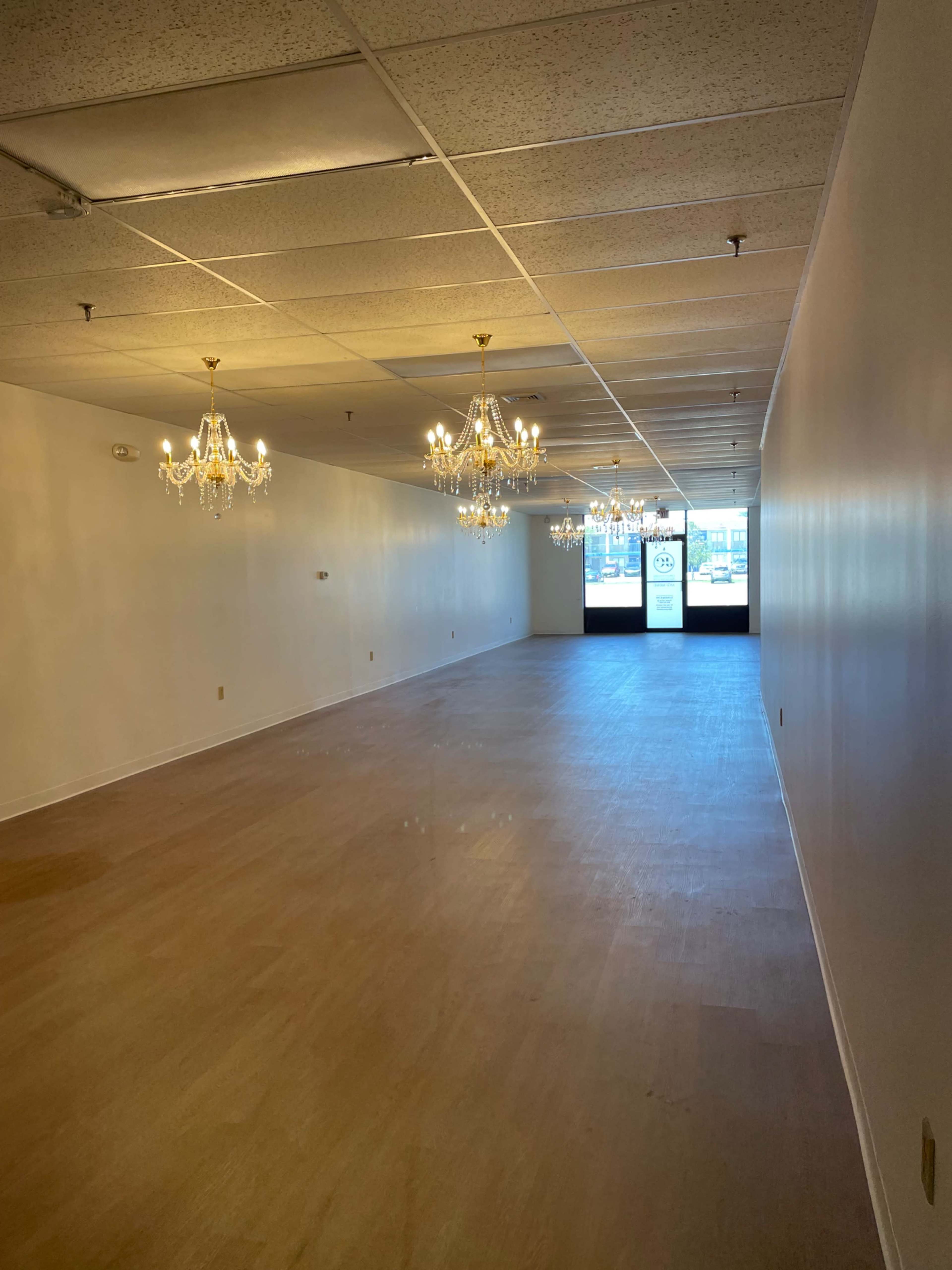 The image shows a spacious, empty retail space with wooden flooring and several chandeliers hanging from the ceiling.