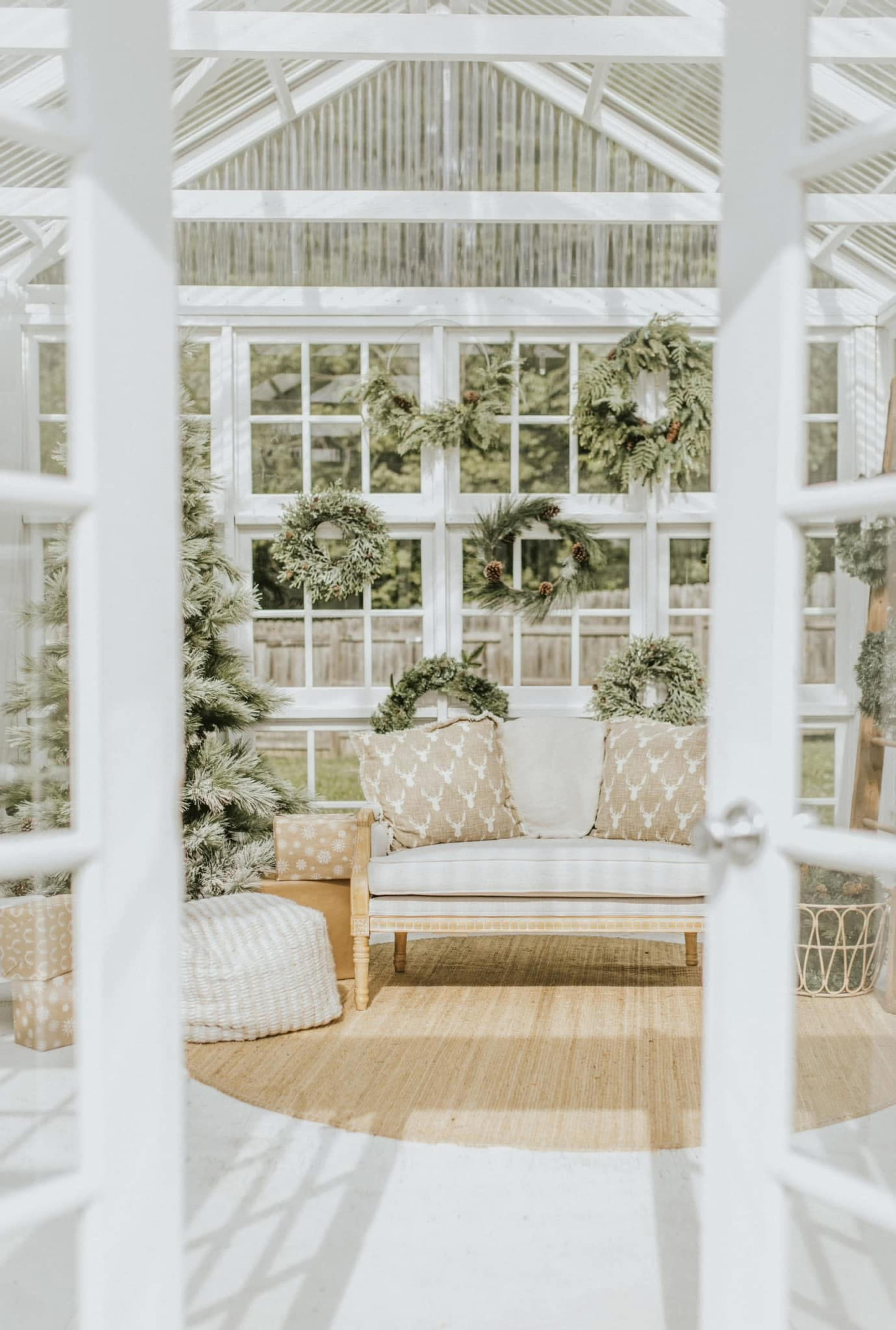 A bright and airy greenhouse features a cozy seating area with two patterned chairs, adorned with wreaths and surrounded by greenery.
