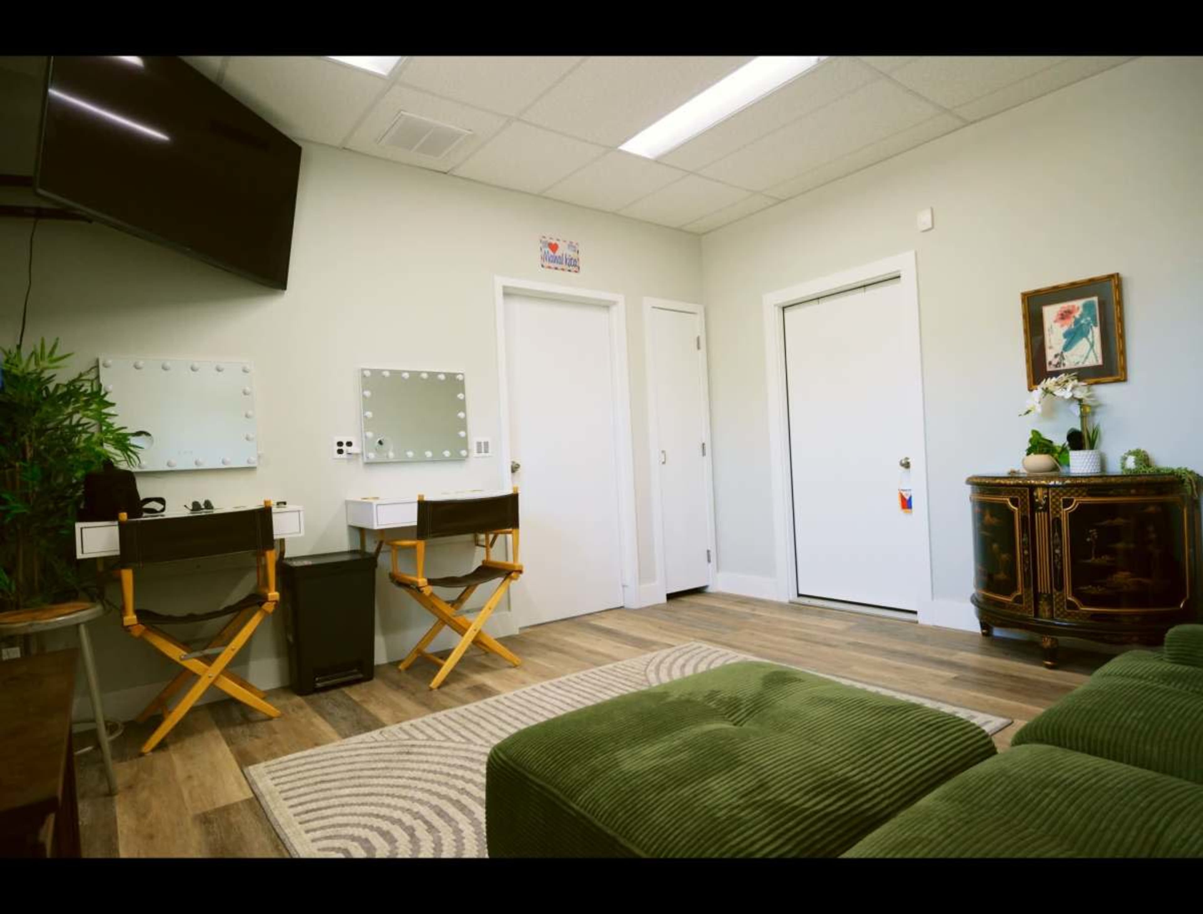 The image shows a small room with two makeup stations, a television mounted on the wall, a green couch, and a closed door leading to another area.