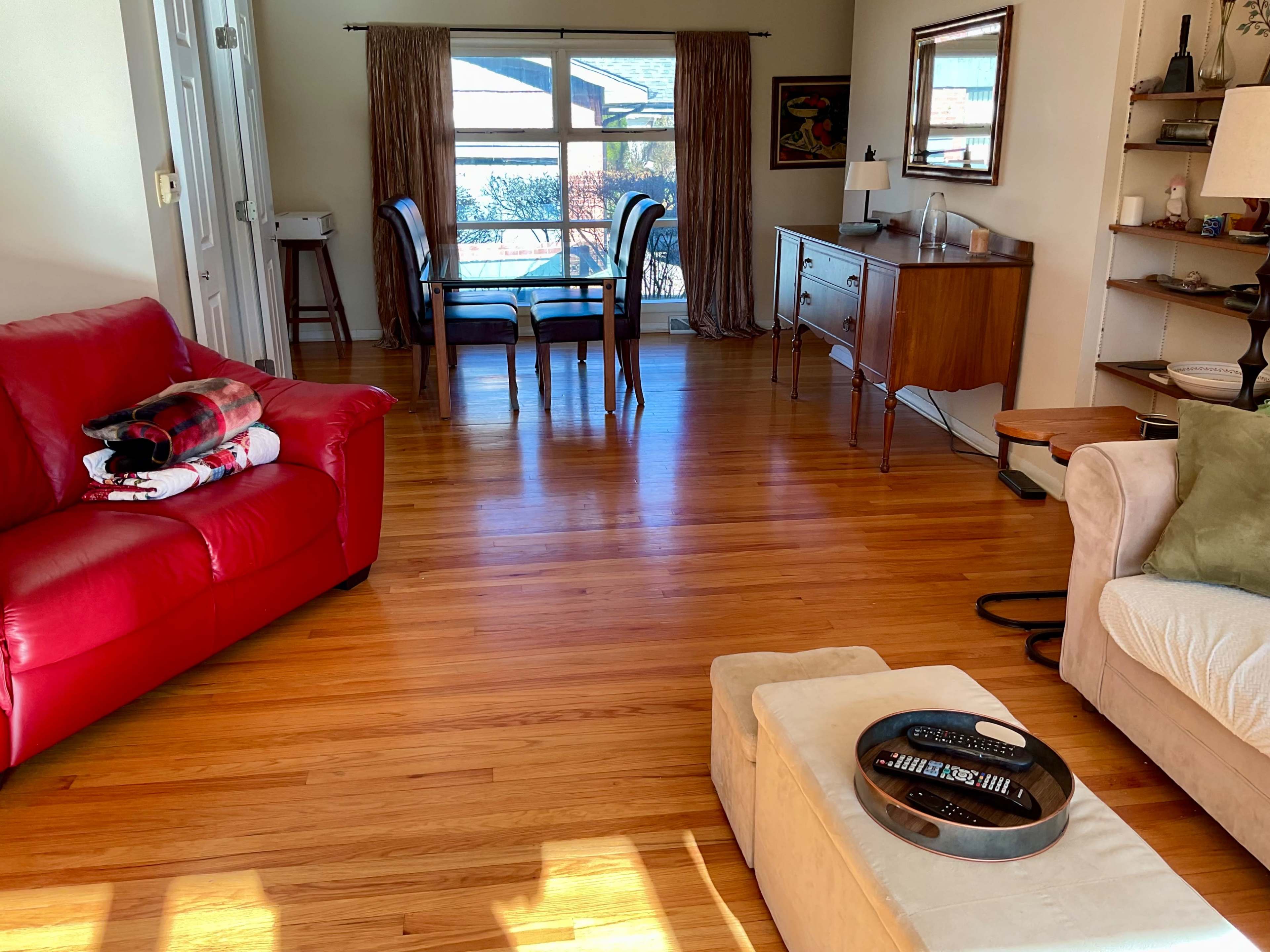 The image shows a living room with hardwood floors, a red sofa, a beige ottoman with a remote on it, a wooden sideboard, and a dining table visible through a large window.