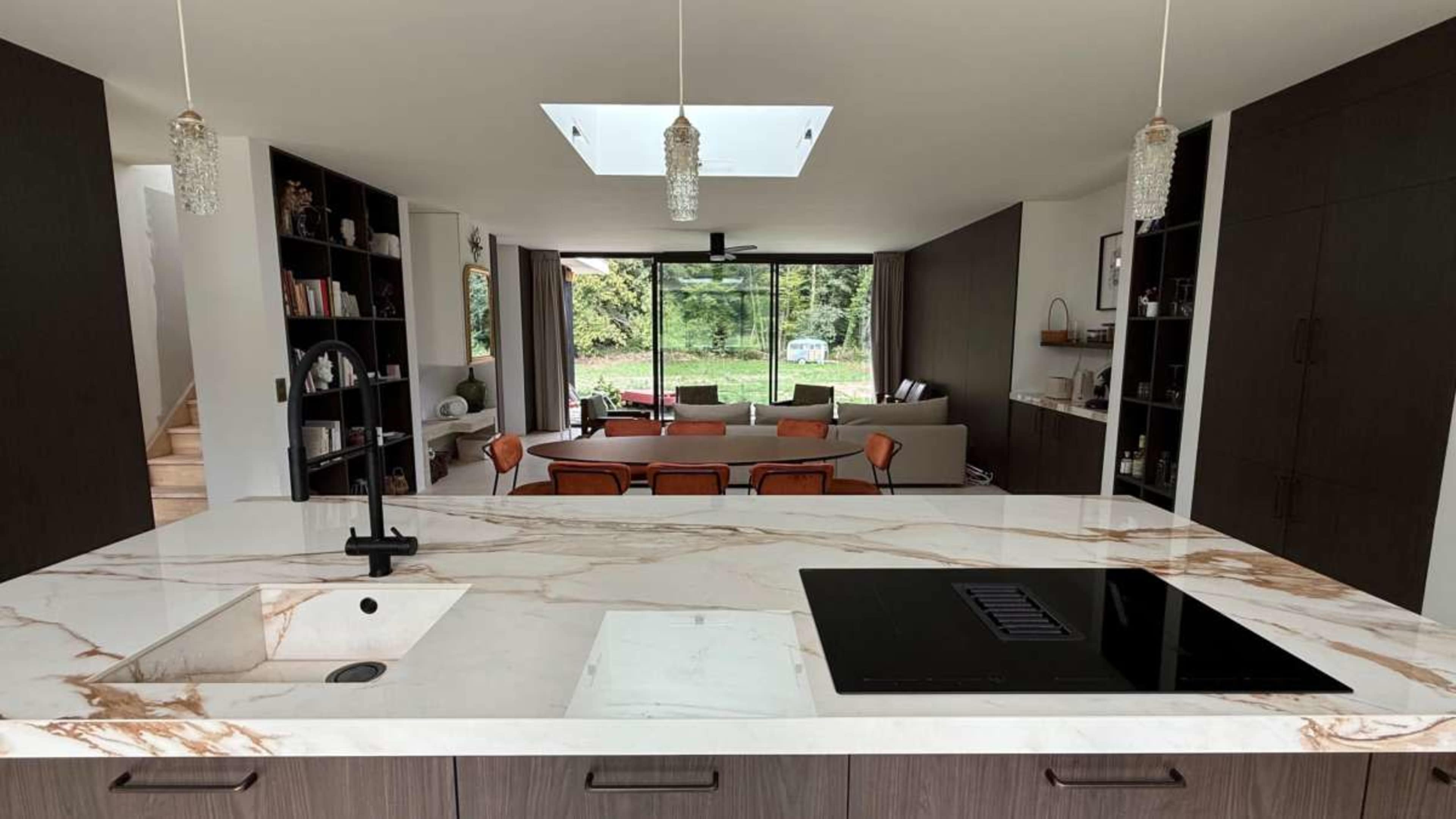 The image shows a modern kitchen with a marble countertop and an open view of a living room that includes a sofa and dining area.